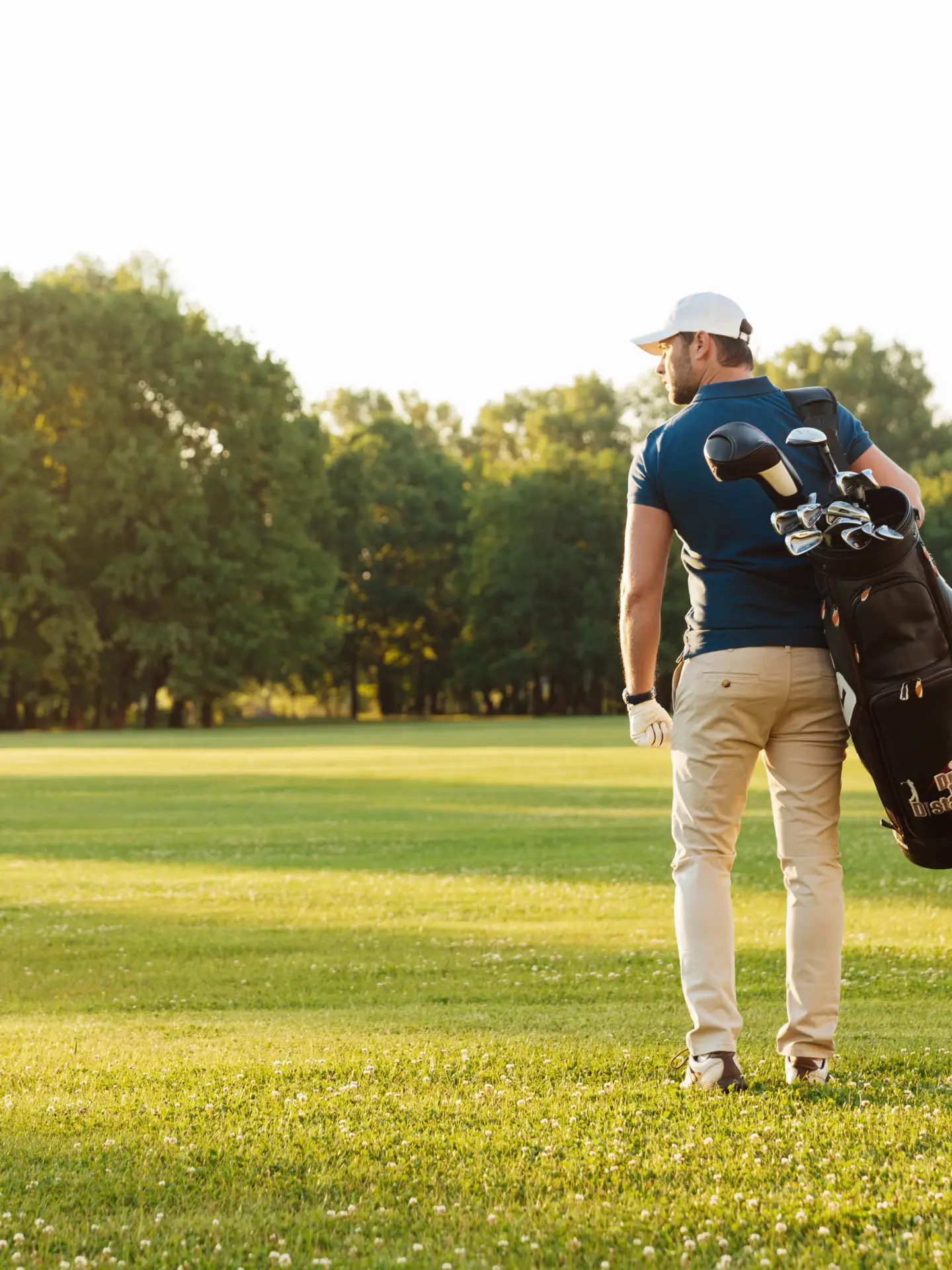 A golfer carrying his golf bag over his shoulder. There are trees in the background and the sun is shining.