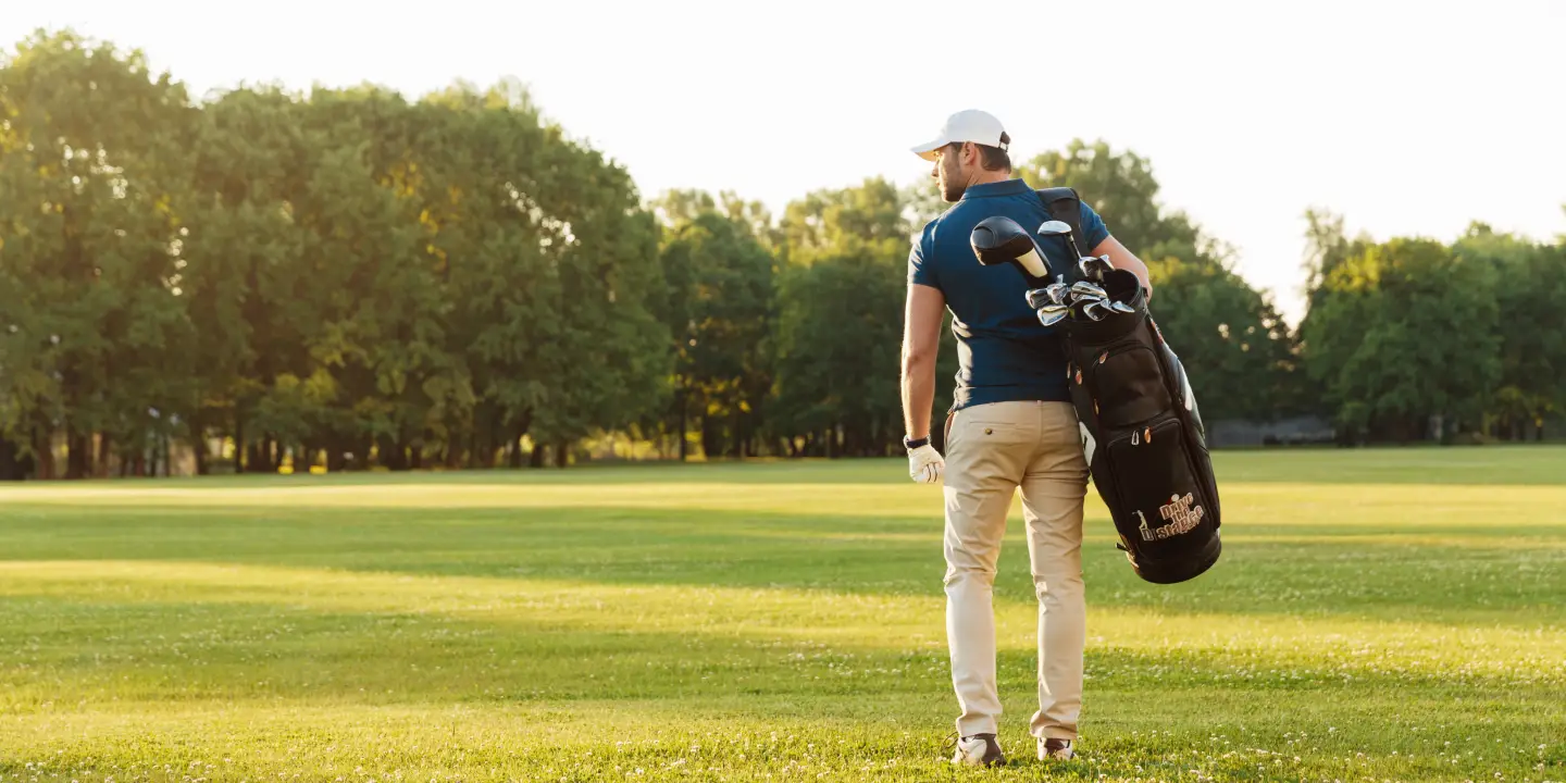 A golfer carrying his golf bag over his shoulder. There are trees in the background and the sun is shining.