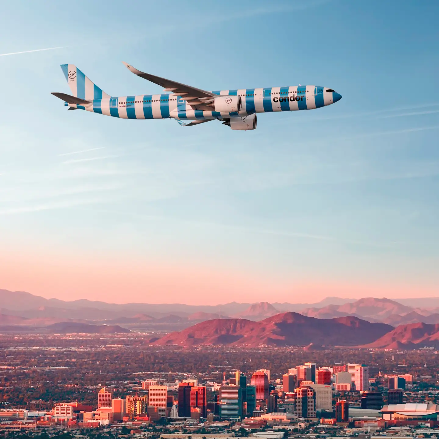 Condor aircraft with blue and white stripes flying over an urban skyline and reddish mountains at dusk.
