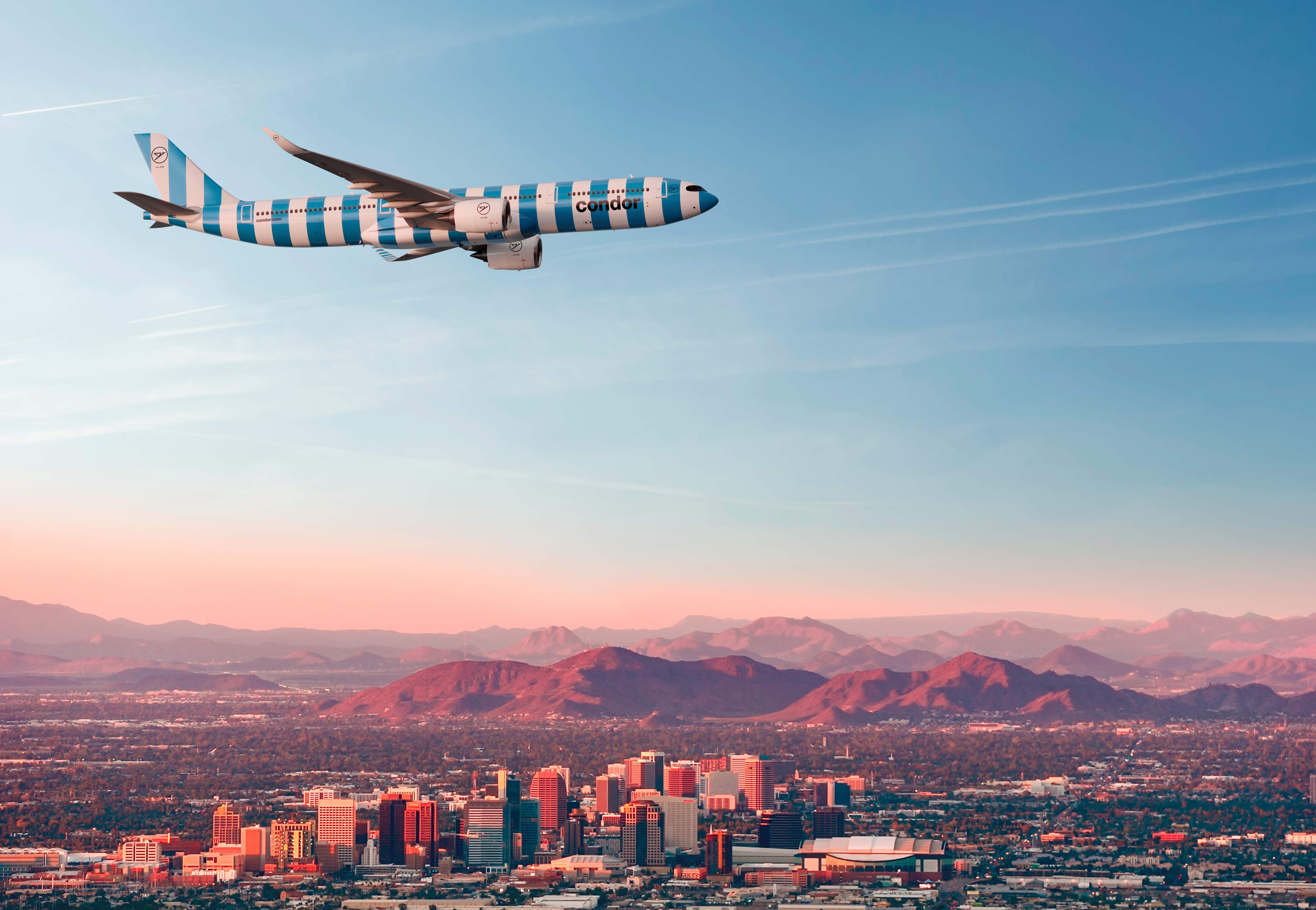 Condor Flugzeug mit blau-weißer Streifenlackierung im Flug über urbaner Skyline und rötlichen Bergen bei Dämmerung.