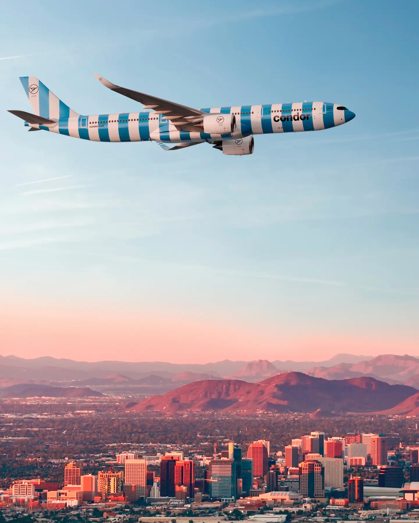 Condor Flugzeug mit blau-weißer Streifenlackierung im Flug über urbaner Skyline und rötlichen Bergen bei Dämmerung.