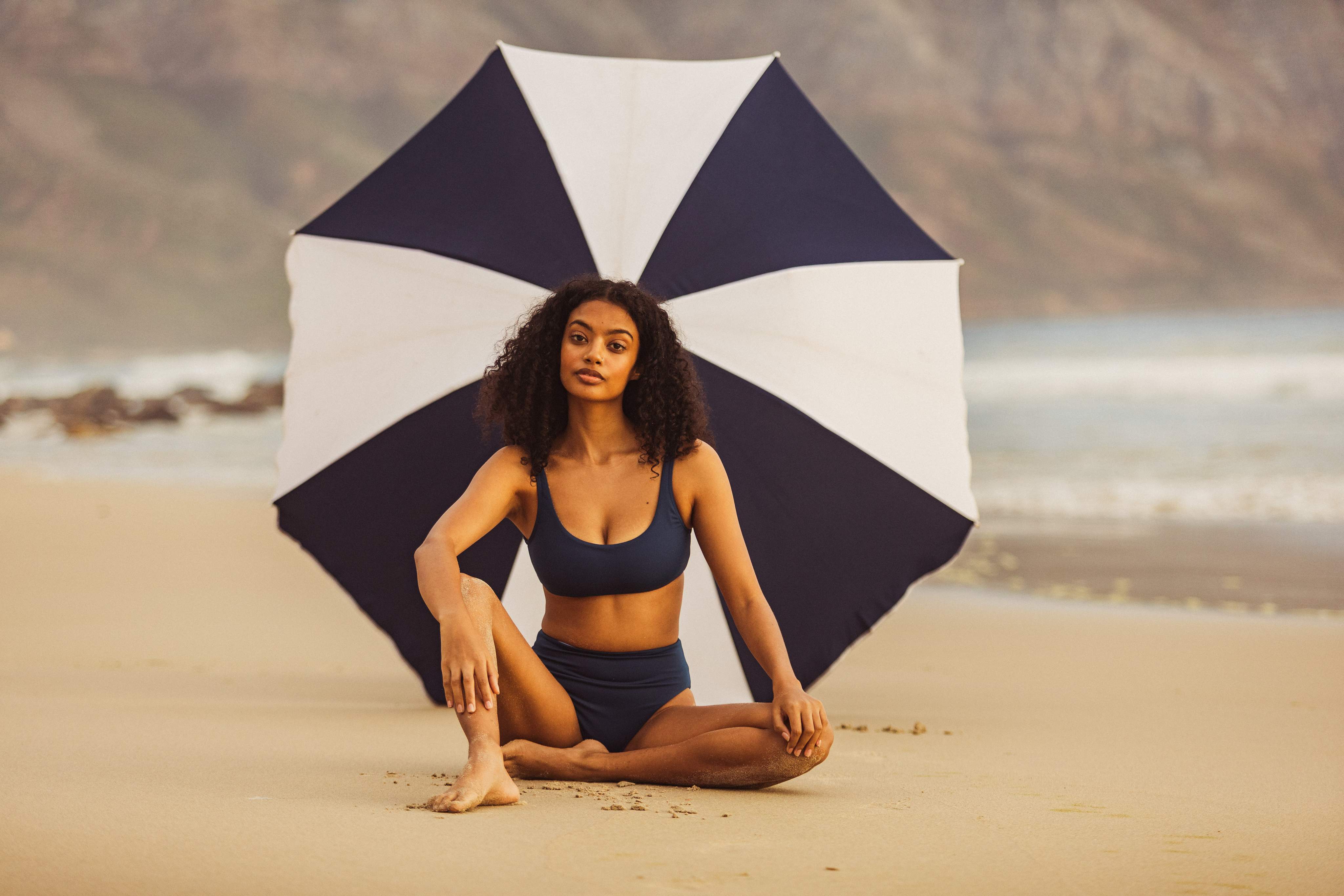 Une femme assise sur la plage sous un parasol rayé bleu et blanc qui la protège du soleil.