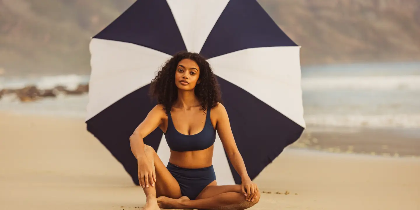 Une femme assise sur la plage sous un parasol rayé bleu et blanc qui la protège du soleil.