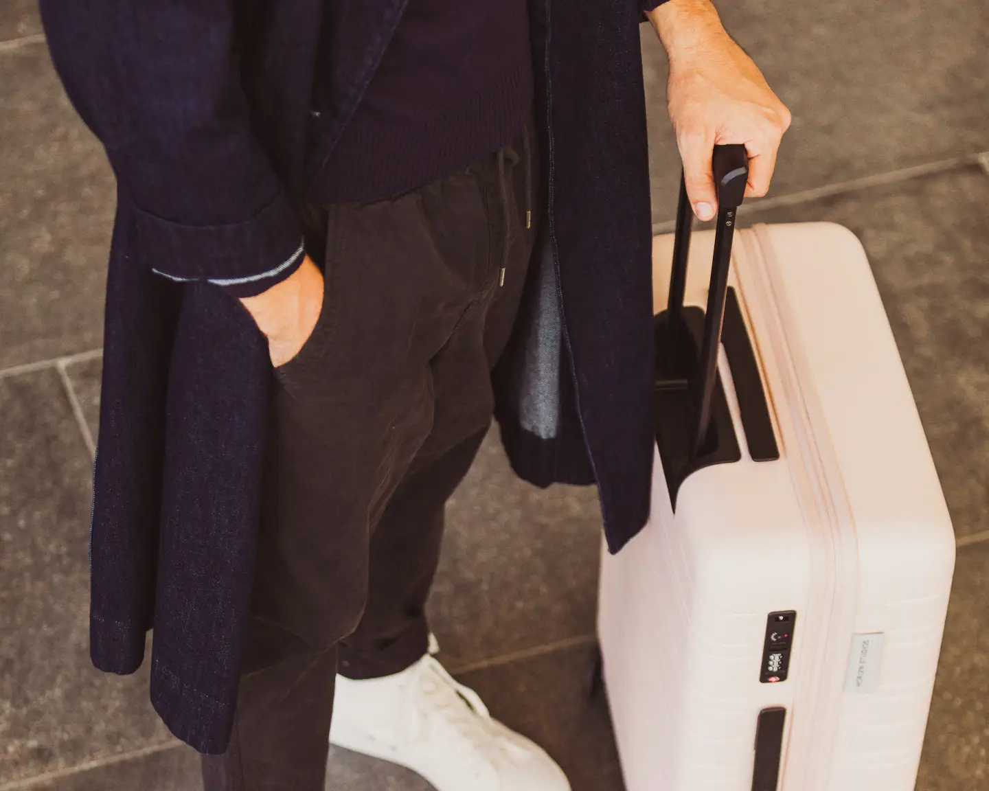 Opname van een man vanaf de schouders naar beneden, die een jas en witte sneakers draagt en een lichtroze koffer vasthoudt.