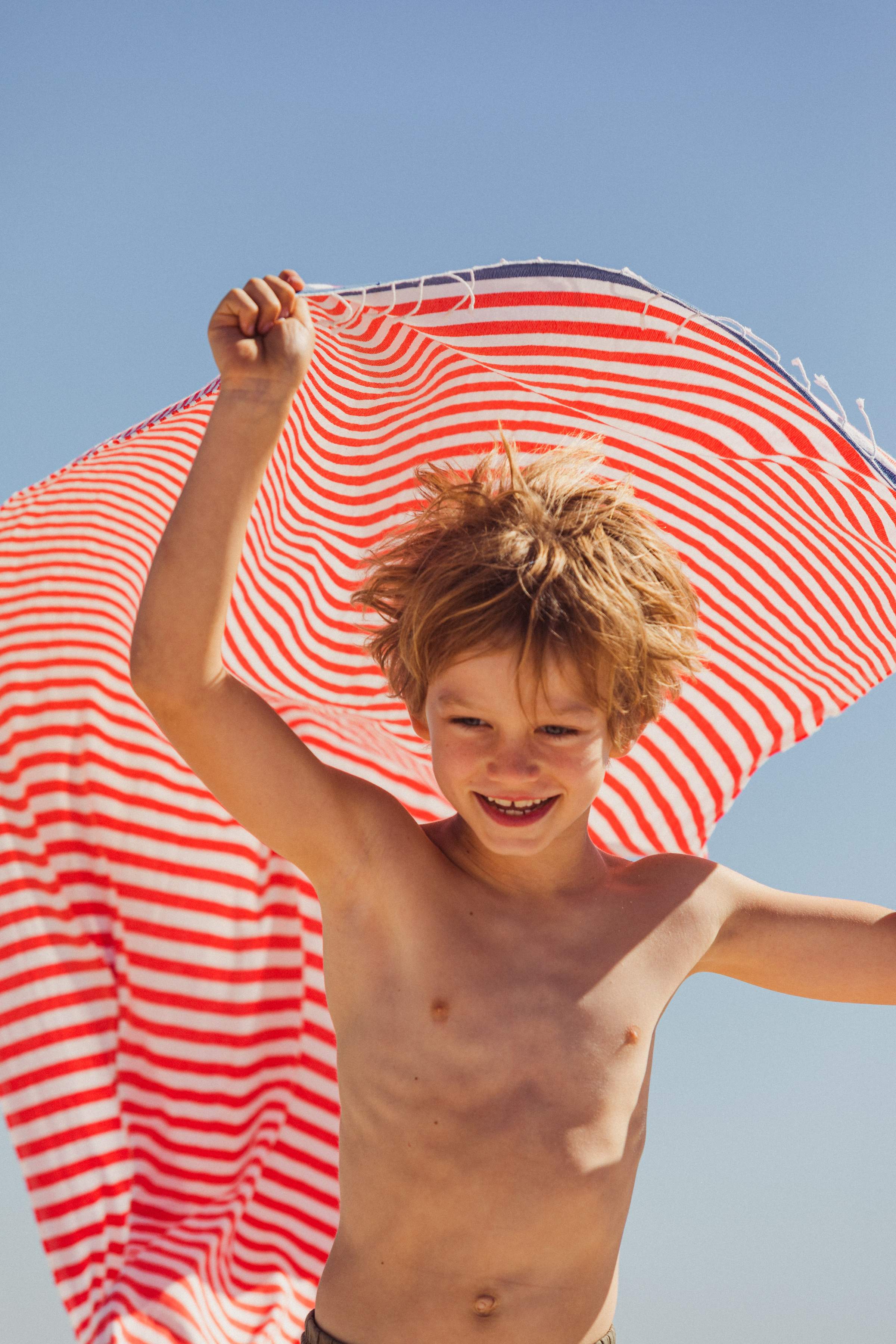 Un niño alegre en la playa sostiene una toalla de rayas rojas y blancas sobre la cabeza.