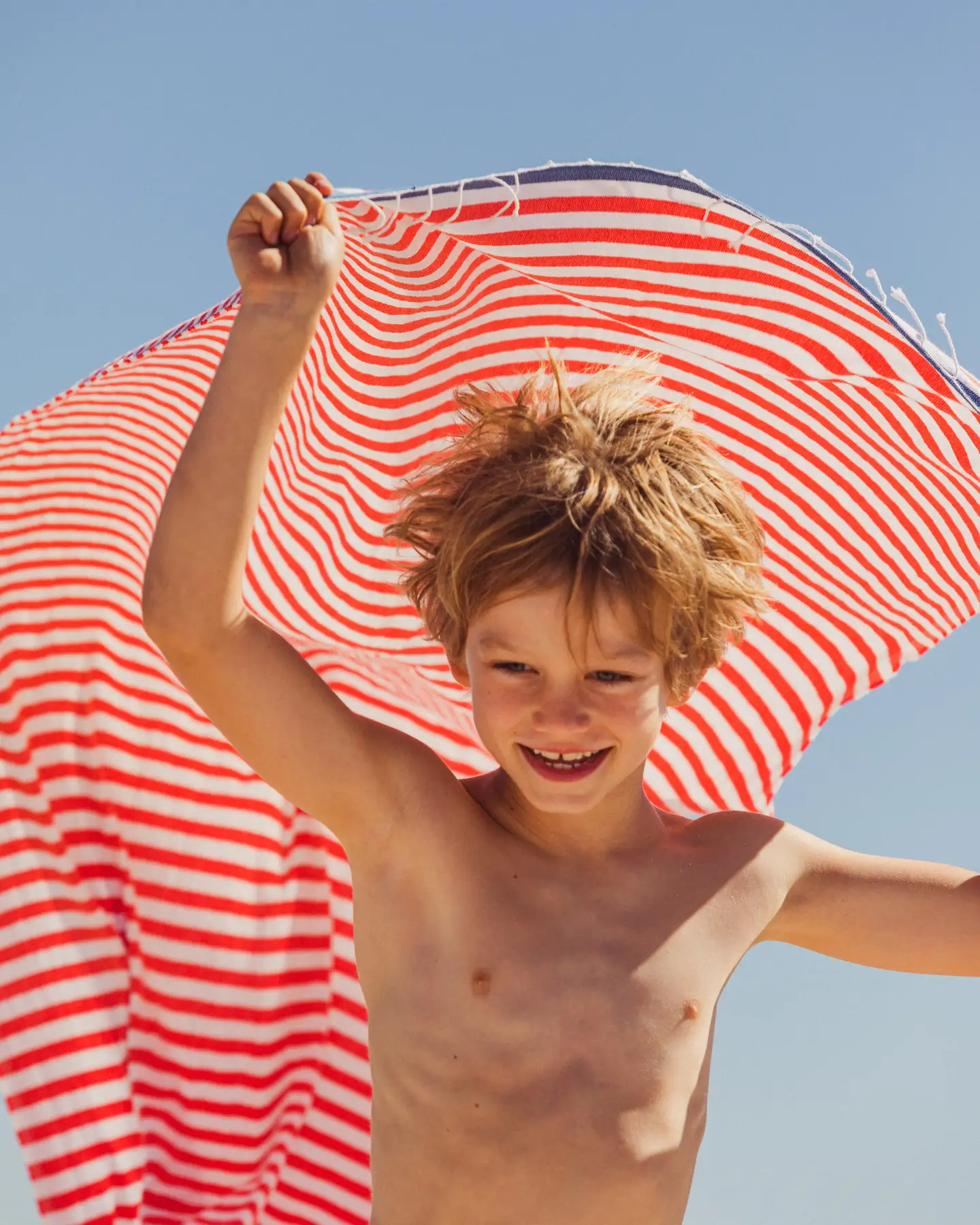 Un niño alegre en la playa sostiene una toalla de rayas rojas y blancas sobre la cabeza.