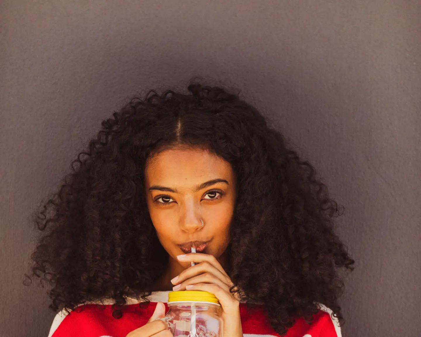 Mujer joven con camiseta de rayas rojas y blancas, cabello rizado y piercing en la nariz, bebiendo de un vaso con tapa amarilla frente a una pared gris.