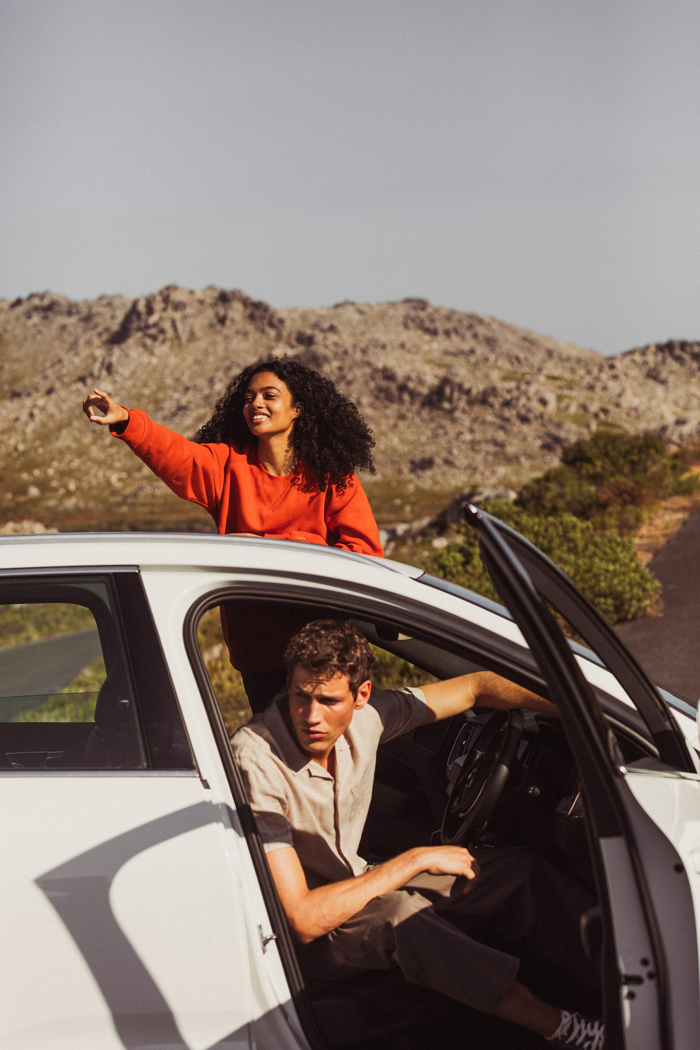 Mujer y hombre de pie junto a su coche de alquiler en una carretera. Se pueden ver rocas escarpadas al fondo y el sol brilla.