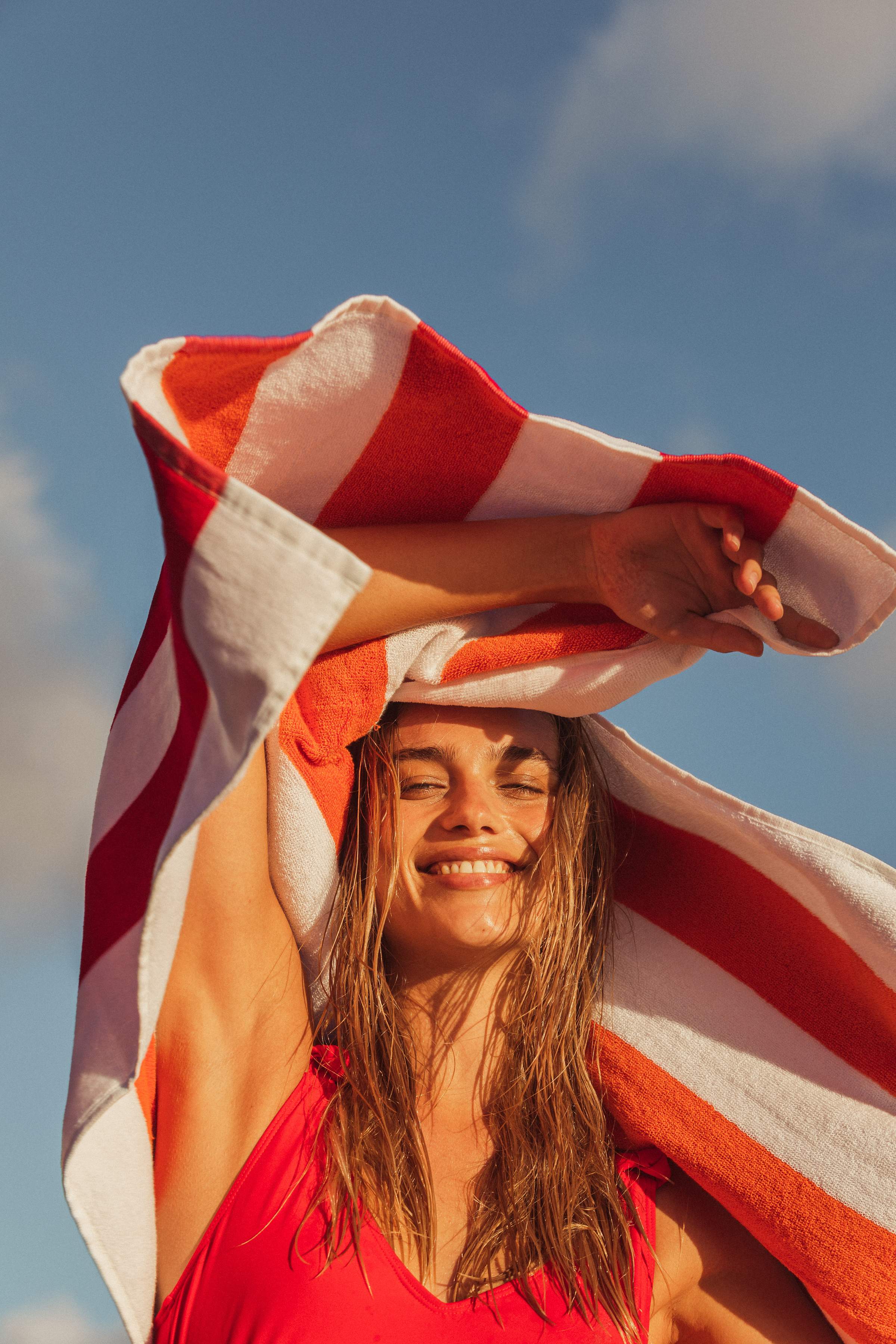 Mujer joven sonriendo y sosteniendo una toalla de playa roja y blanca sobre la cabeza, contra un cielo soleado.