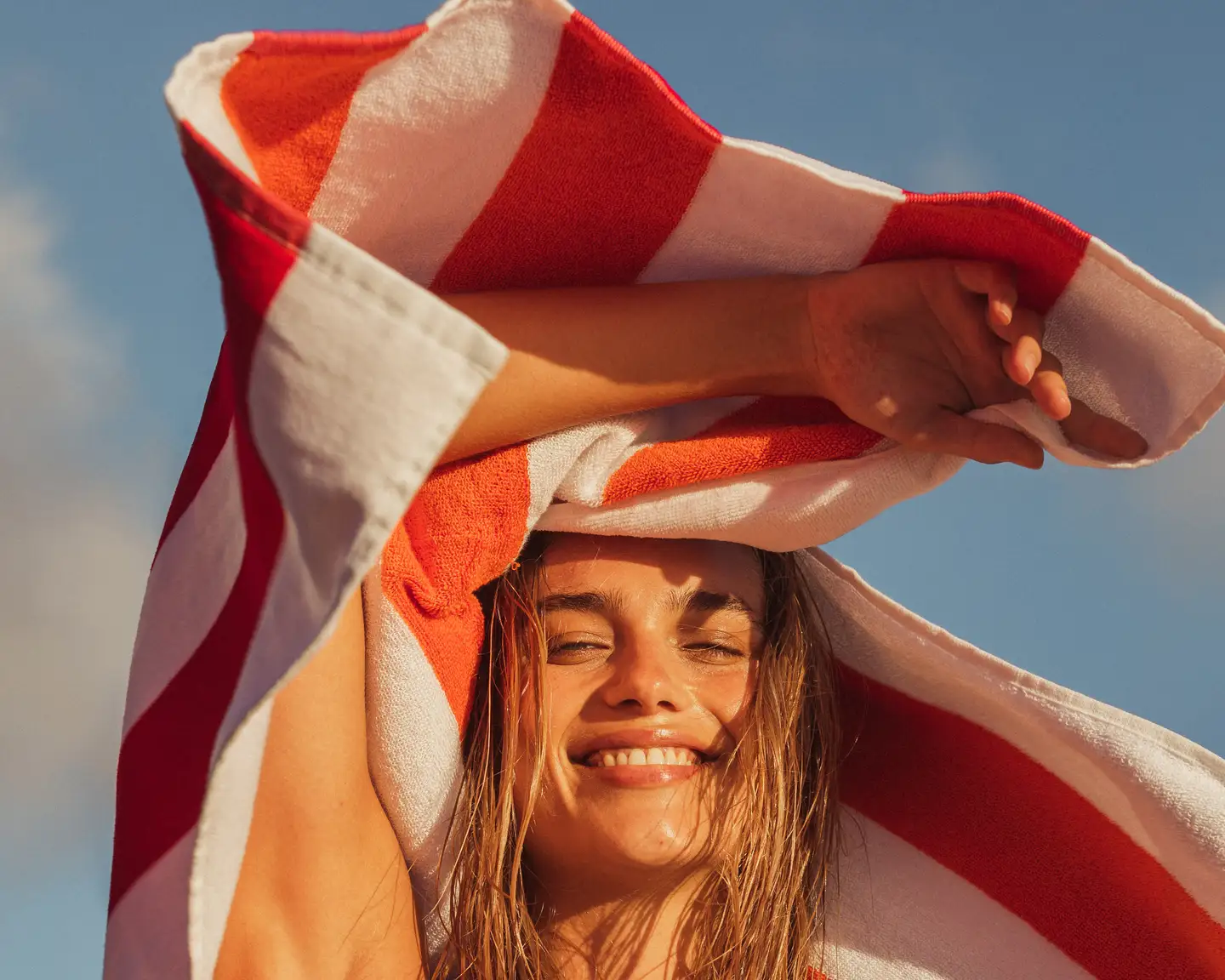 Mujer joven sonriendo y sosteniendo una toalla de playa roja y blanca sobre la cabeza, contra un cielo soleado.