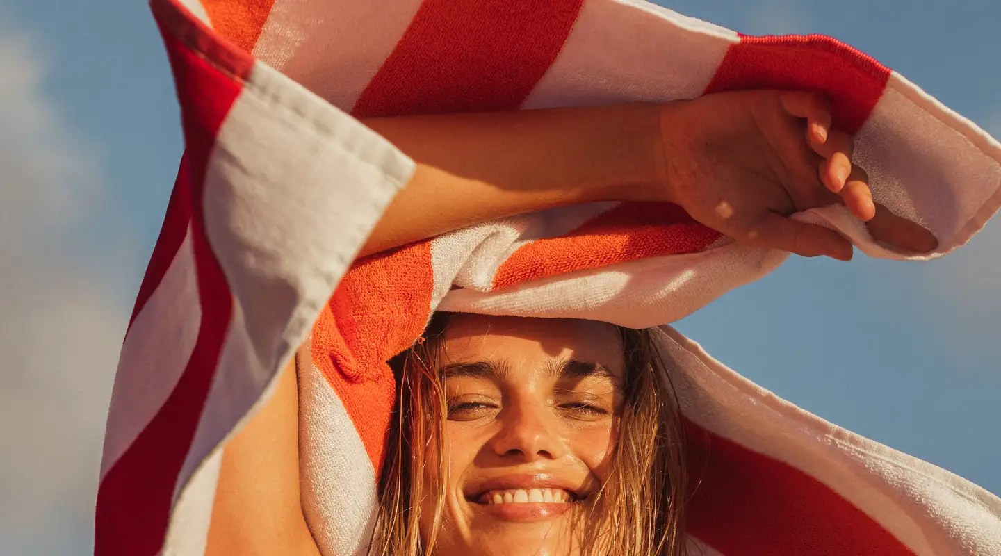 Mujer joven sonriendo y sosteniendo una toalla de playa roja y blanca sobre la cabeza, contra un cielo soleado.