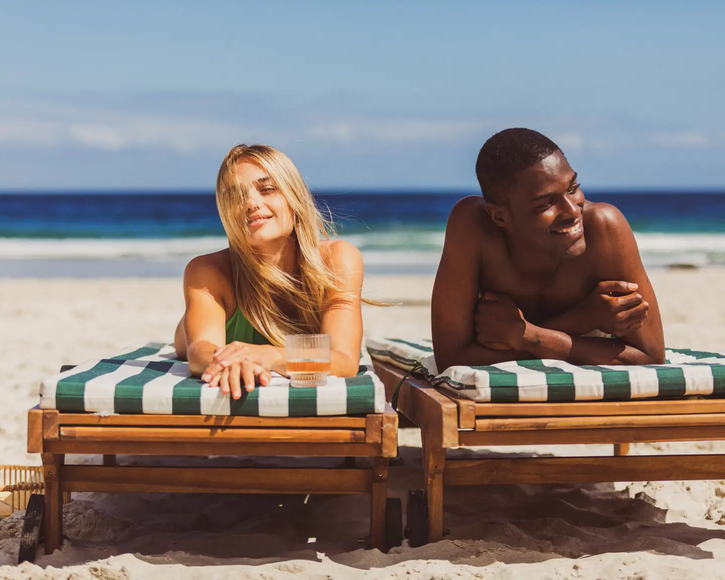 Ein junger Mann und eine junge Frau lächeln und unterhalten sich, während sie auf grün-weiß-gestreiften Liegestühlen am sonnigen Strand entspannen.