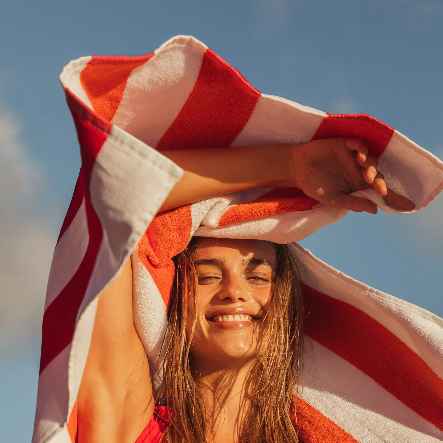 Mujer joven sonriendo y sosteniendo una toalla de playa roja y blanca sobre la cabeza contra un cielo soleado.