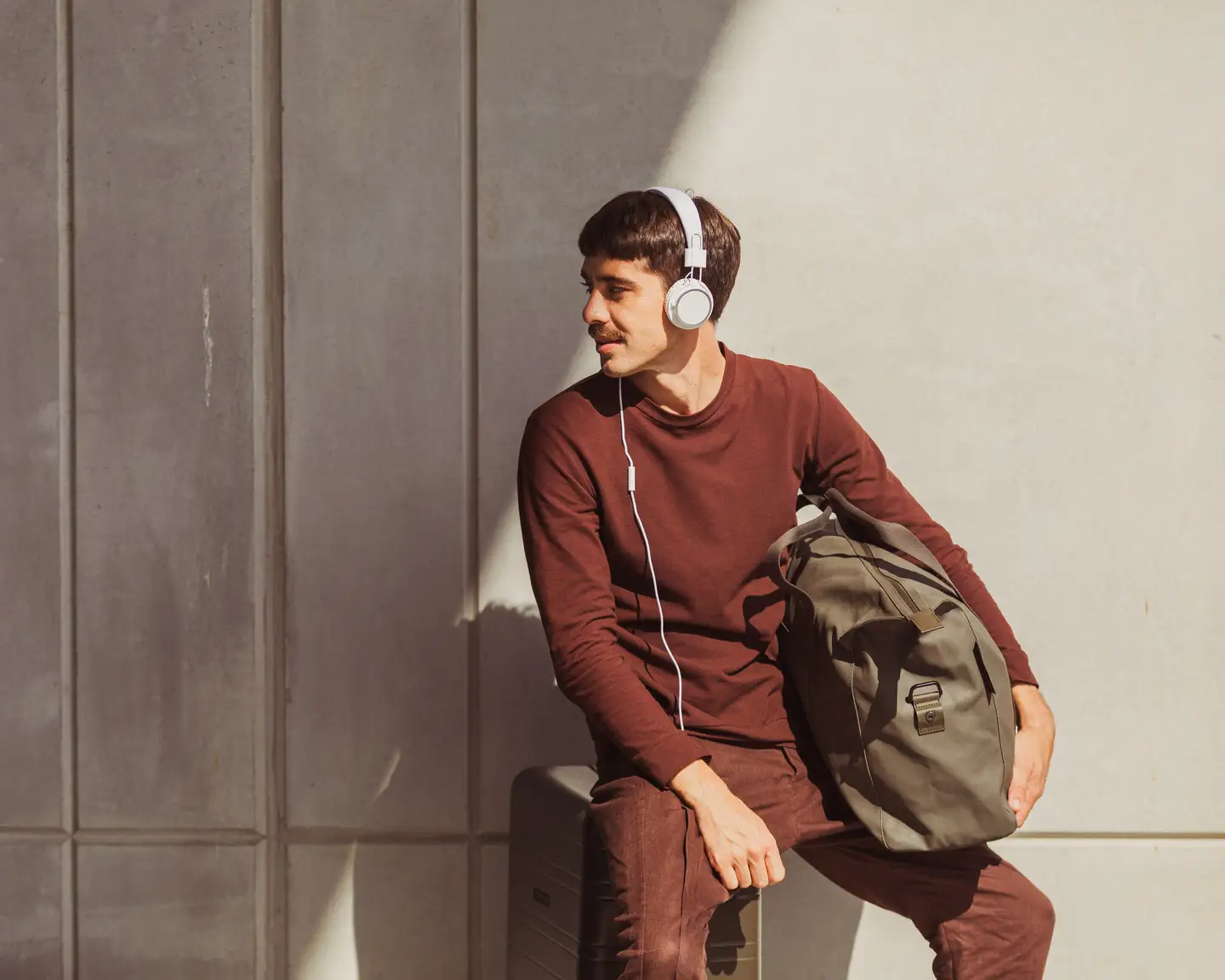 Un hombre se sienta en una maleta con los auriculares puestos, sosteniendo su bolsa de viaje