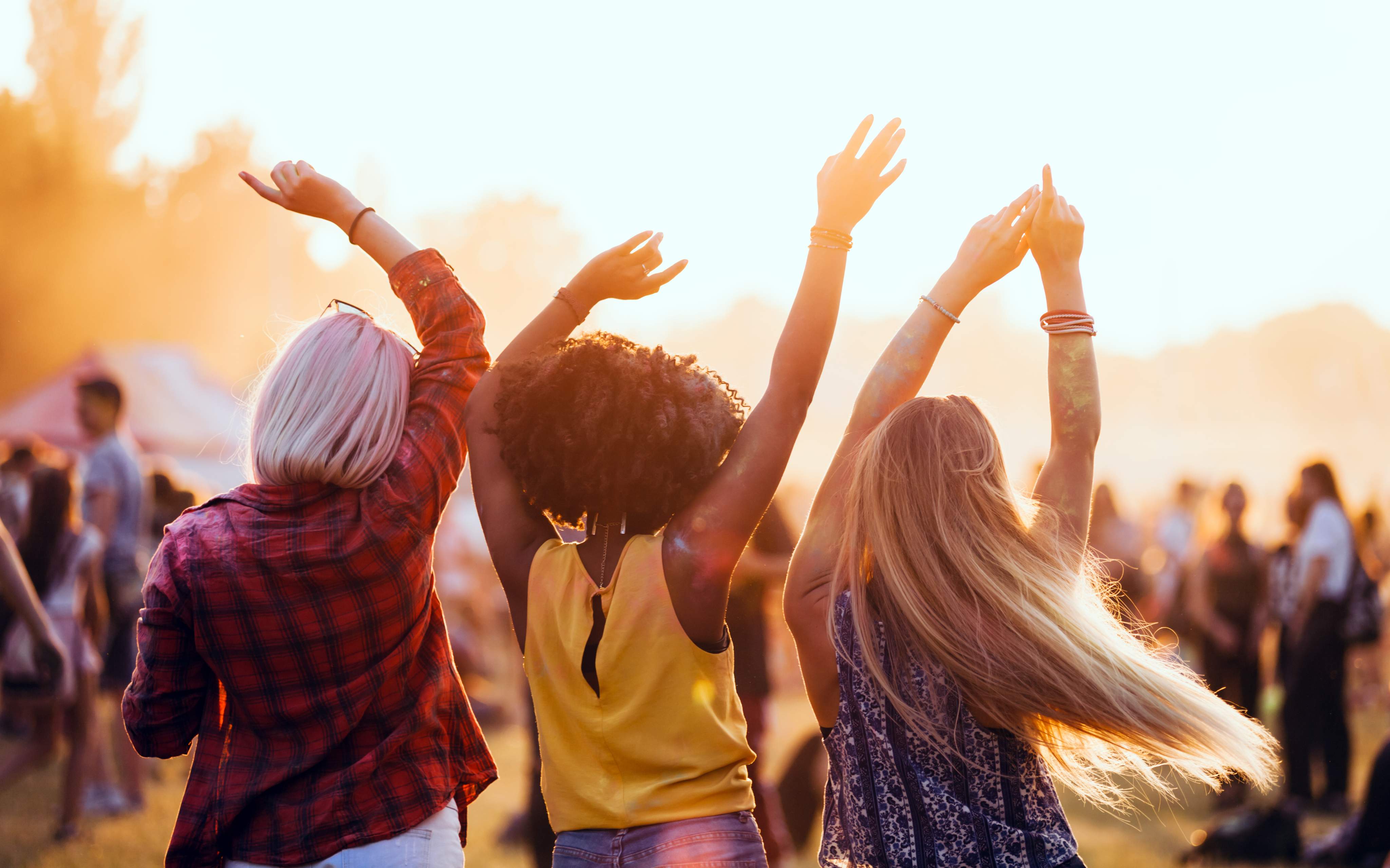 Drei Freunde genießen ein Open-Air-Festival und tanzen mit erhobenen Armen in der Abendsonne.