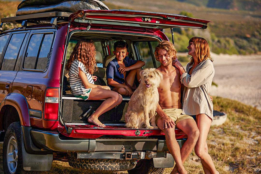 Uma família na praia com um grande carro vermelho. Duas crianças e um homem estão sentados juntos com um cão no porta-malas do carro, a mulher está ao lado deles.