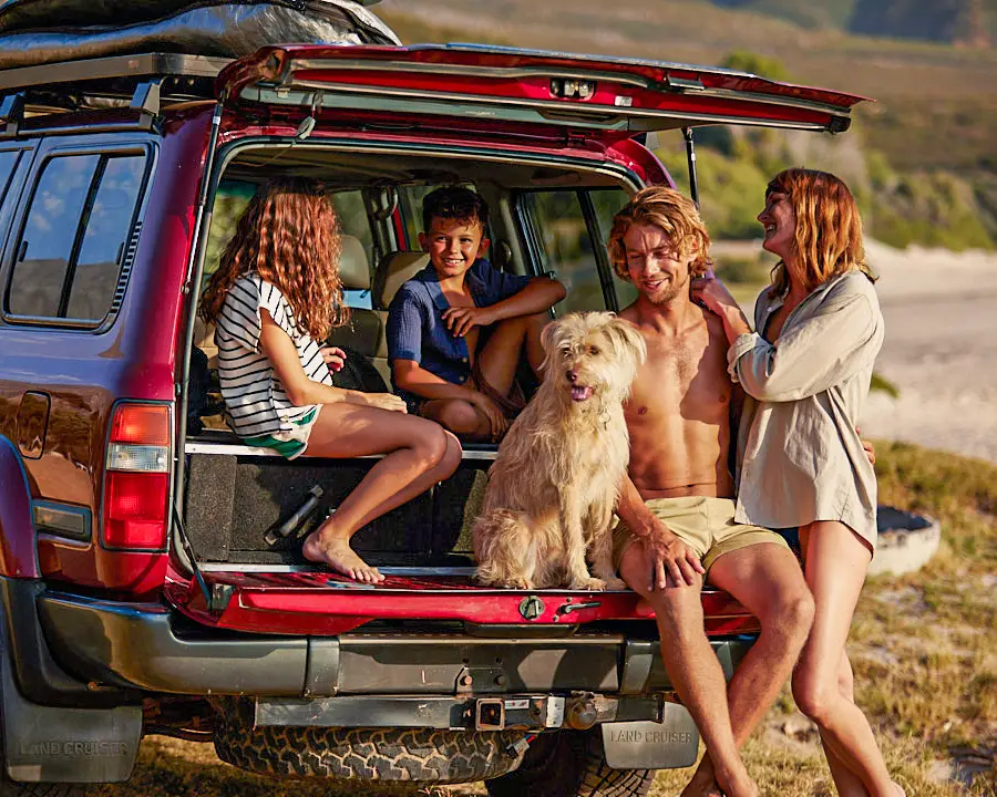 Uma família na praia com um grande carro vermelho. Duas crianças e um homem estão sentados juntos com um cão no porta-malas do carro, a mulher está ao lado deles.
