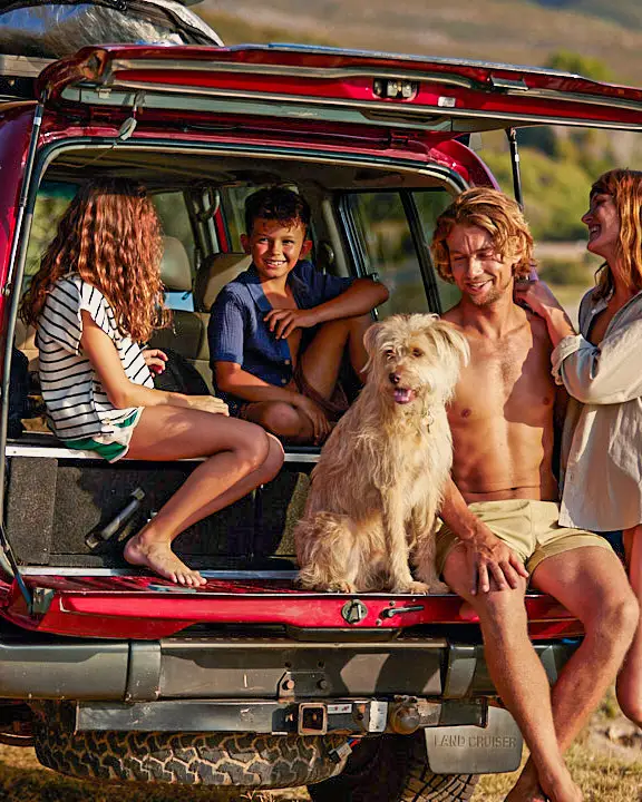 Uma família na praia com um grande carro vermelho. Duas crianças e um homem estão sentados juntos com um cão no porta-malas do carro, a mulher está ao lado deles.