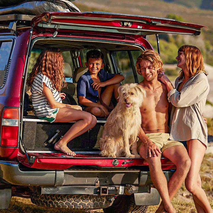 Uma família na praia com um grande carro vermelho. Duas crianças e um homem estão sentados juntos com um cão no porta-malas do carro, a mulher está ao lado deles.