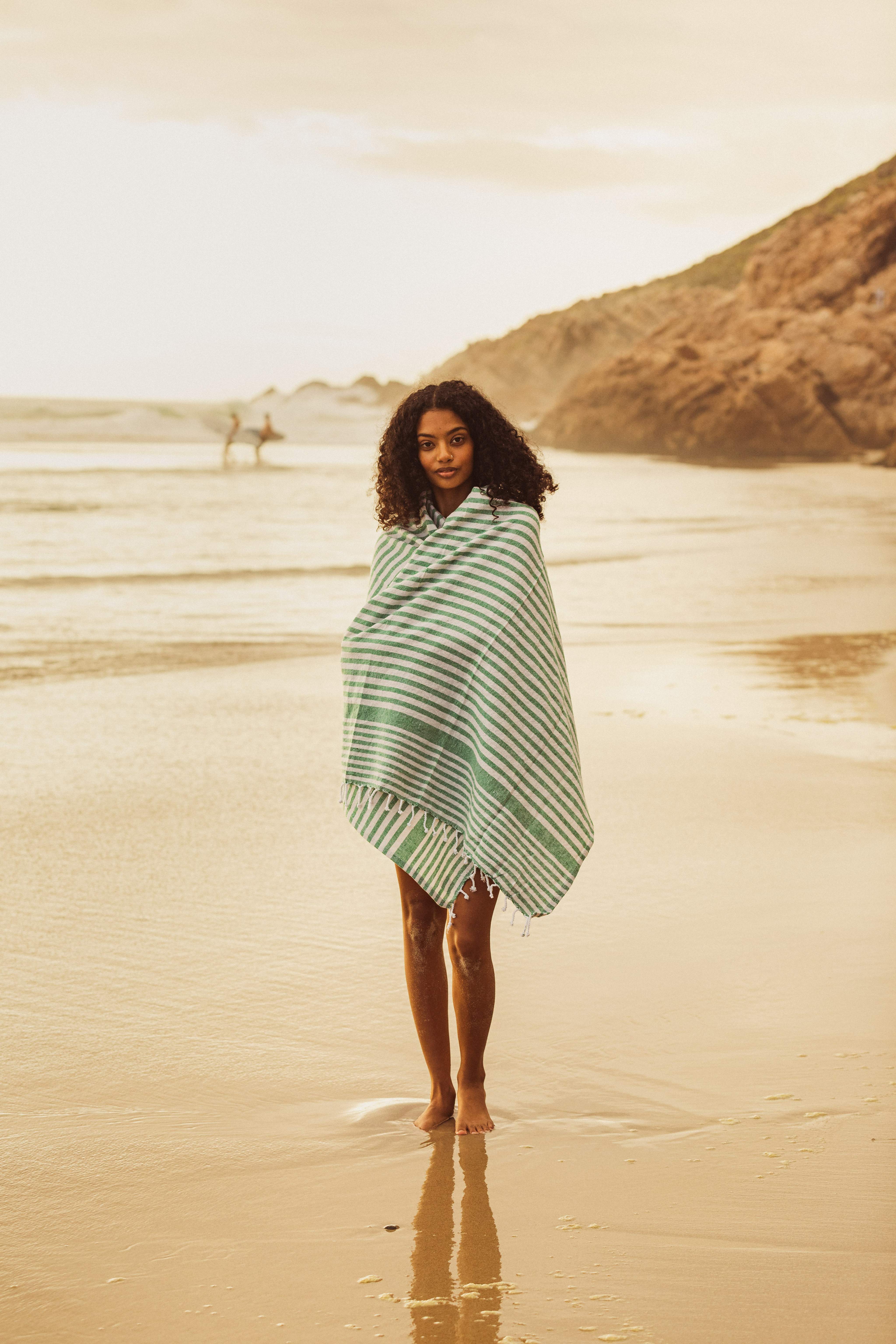 Une femme enveloppée dans une serviette à rayures vertes et blanches prend une photo, debout sur une plage de sable