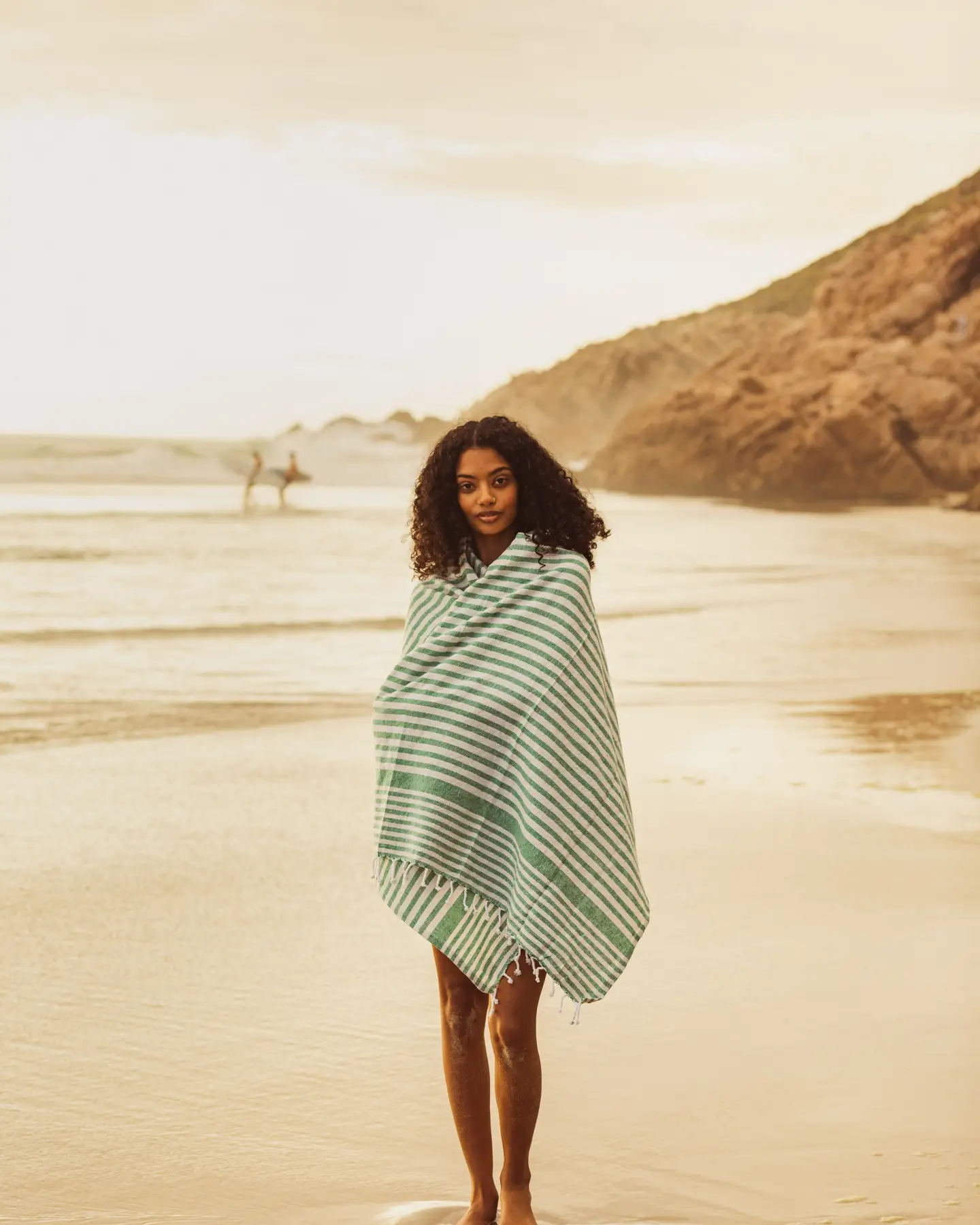 Une femme enveloppée dans une serviette à rayures vertes et blanches prend une photo, debout sur une plage de sable