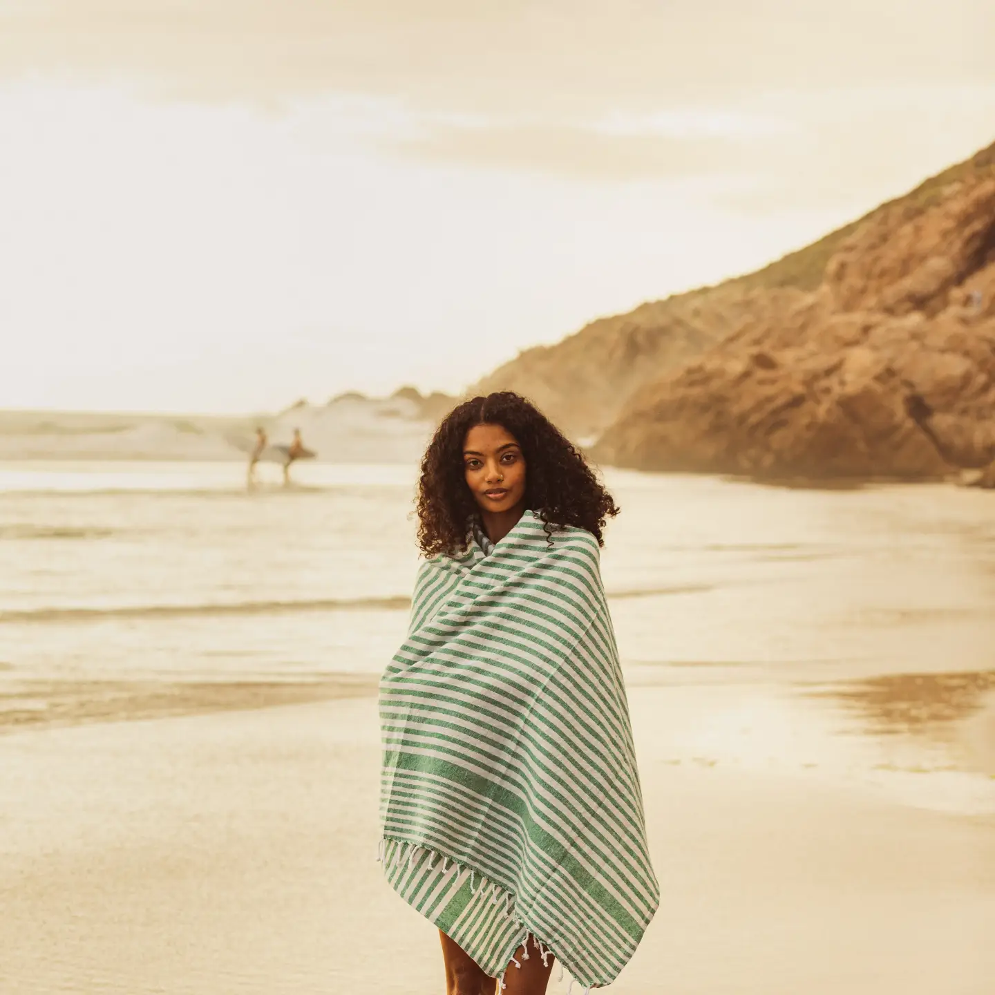 Une femme enveloppée dans une serviette à rayures vertes et blanches prend une photo, debout sur une plage de sable