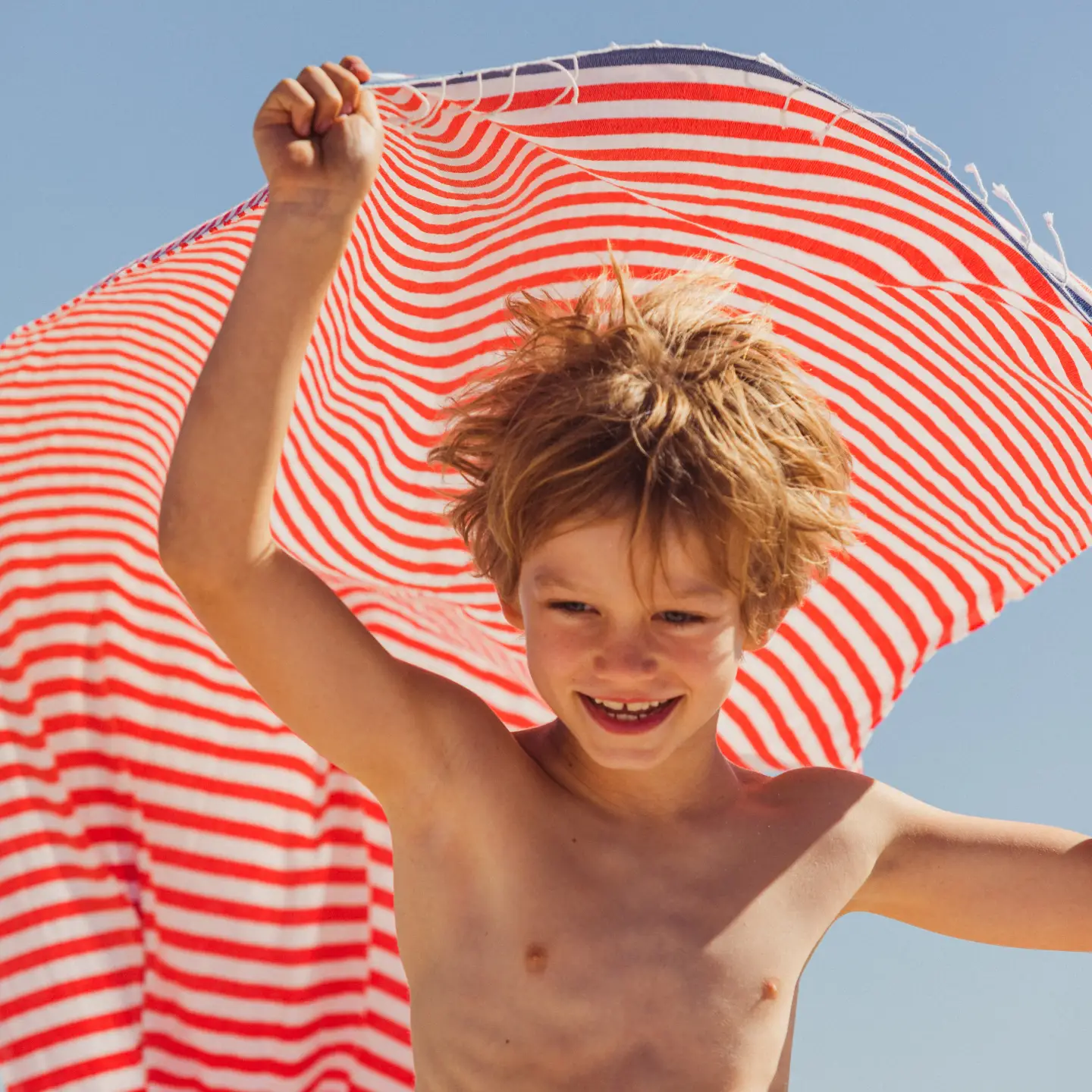 Un garçon joyeux sur la plage tient une serviette de plage rayée rouge et blanche au-dessus de sa tête