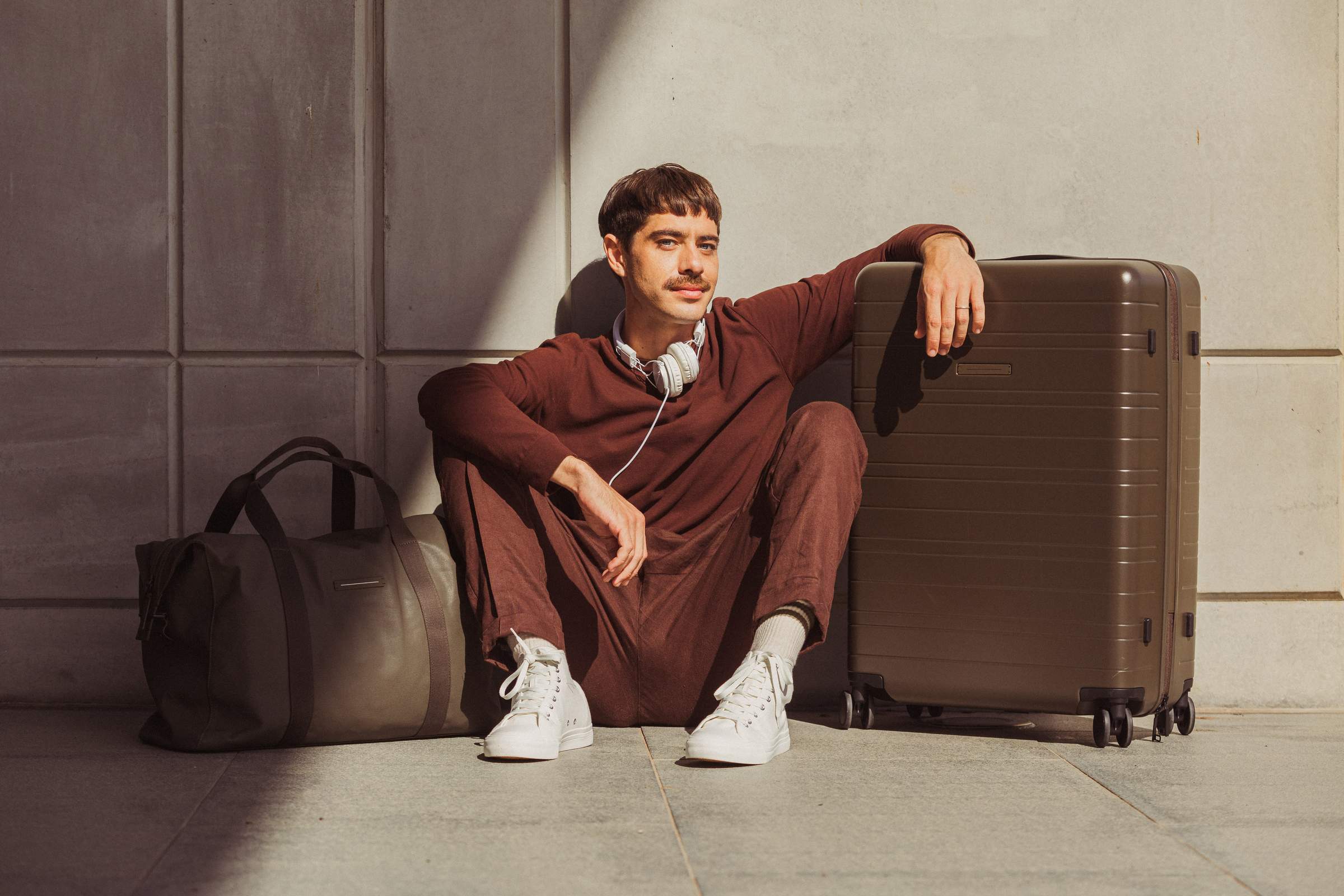 Un hombre de aspecto relajado está sentado junto a una maleta marrón y lleva un traje burdeos y zapatillas blancas. Con los auriculares alrededor del cuello, parece relajado y listo para viajar.