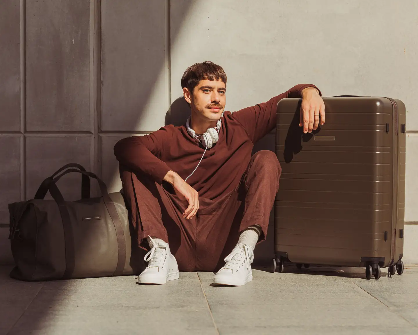 Un hombre de aspecto relajado está sentado junto a una maleta marrón y lleva un traje burdeos y zapatillas blancas. Con los auriculares alrededor del cuello, parece relajado y listo para viajar.