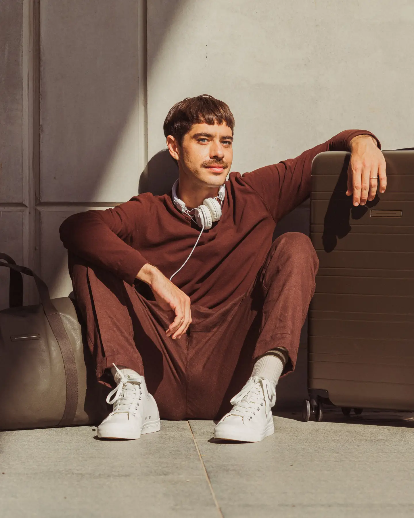 Un homme à l'allure décontractée est assis à côté d'une valise marron. Il porte un vêtement rouge vin et des baskets blanches. Avec des écouteurs autour du cou, il a l'air détendu et prêt à voyager.