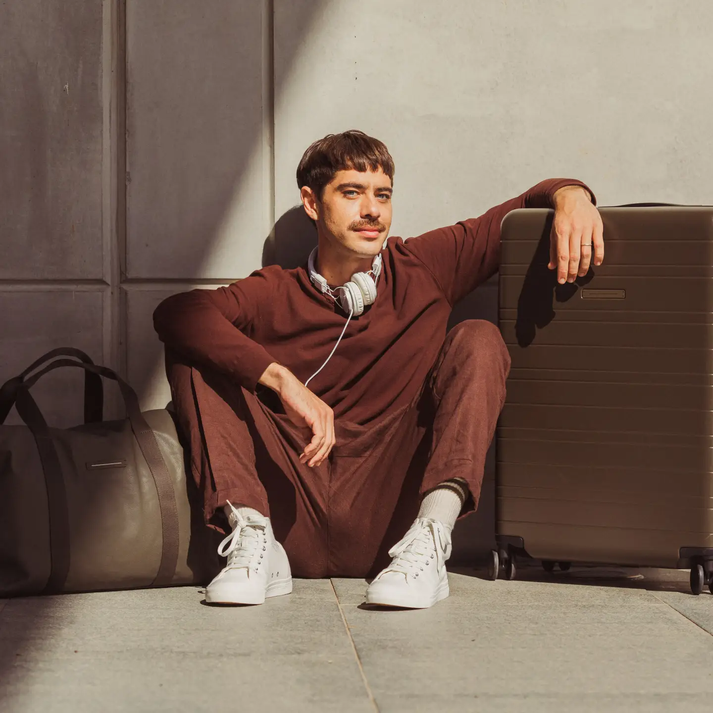 Un homme à l'allure décontractée est assis à côté d'une valise marron. Il porte un vêtement rouge vin et des baskets blanches. Avec des écouteurs autour du cou, il a l'air détendu et prêt à voyager.