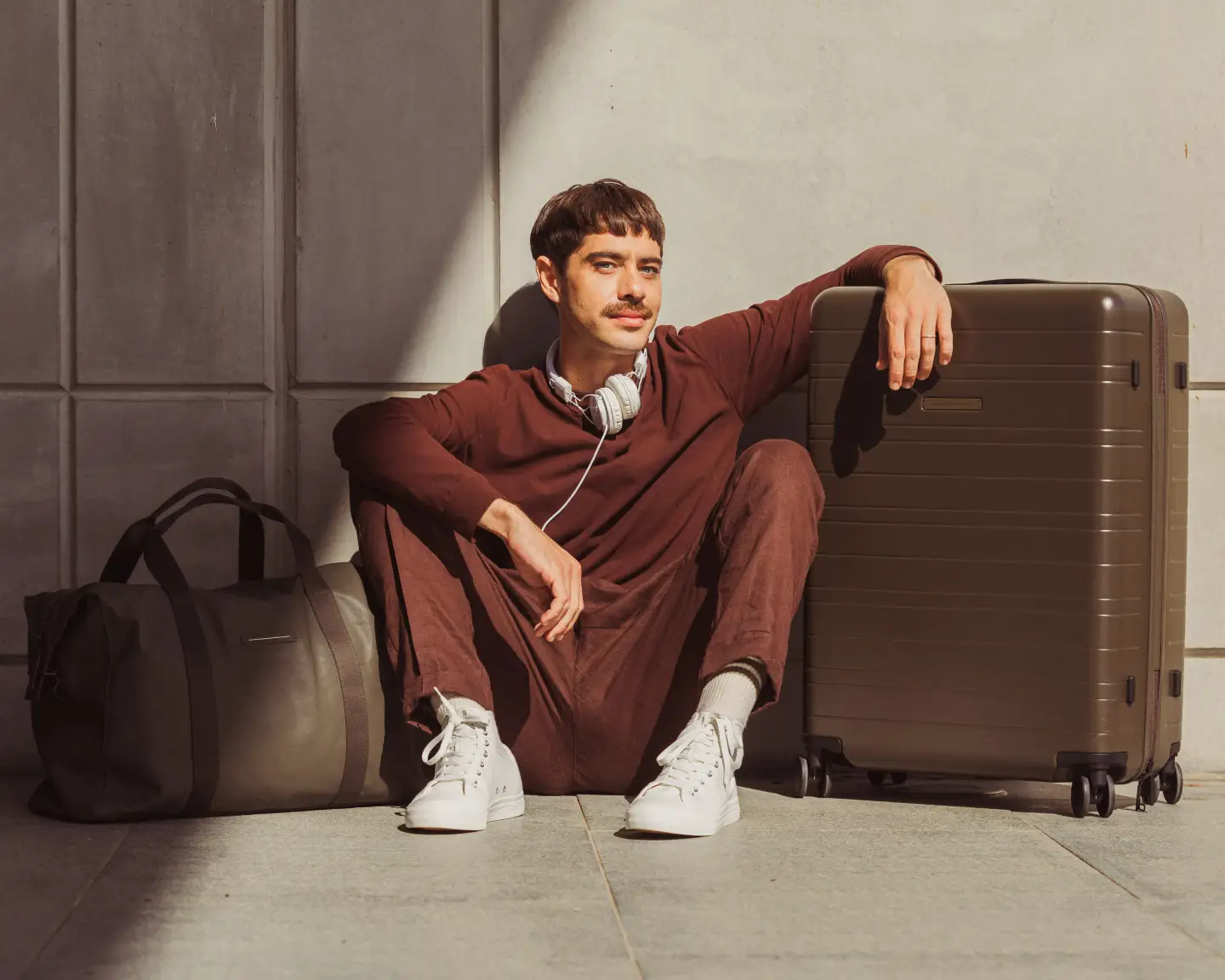 Een ontspannen man in een wijnrode outfit met witte sportschoenen die naast een bruine koffer zit. Met zijn koptelefoon om ziet hij er ontspannen uit en is klaar om te reizen.