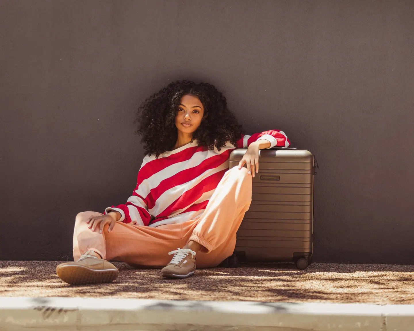 Mujer con un jersey de rayas y pantalones de color claro sentada junto a una maleta pequeña.