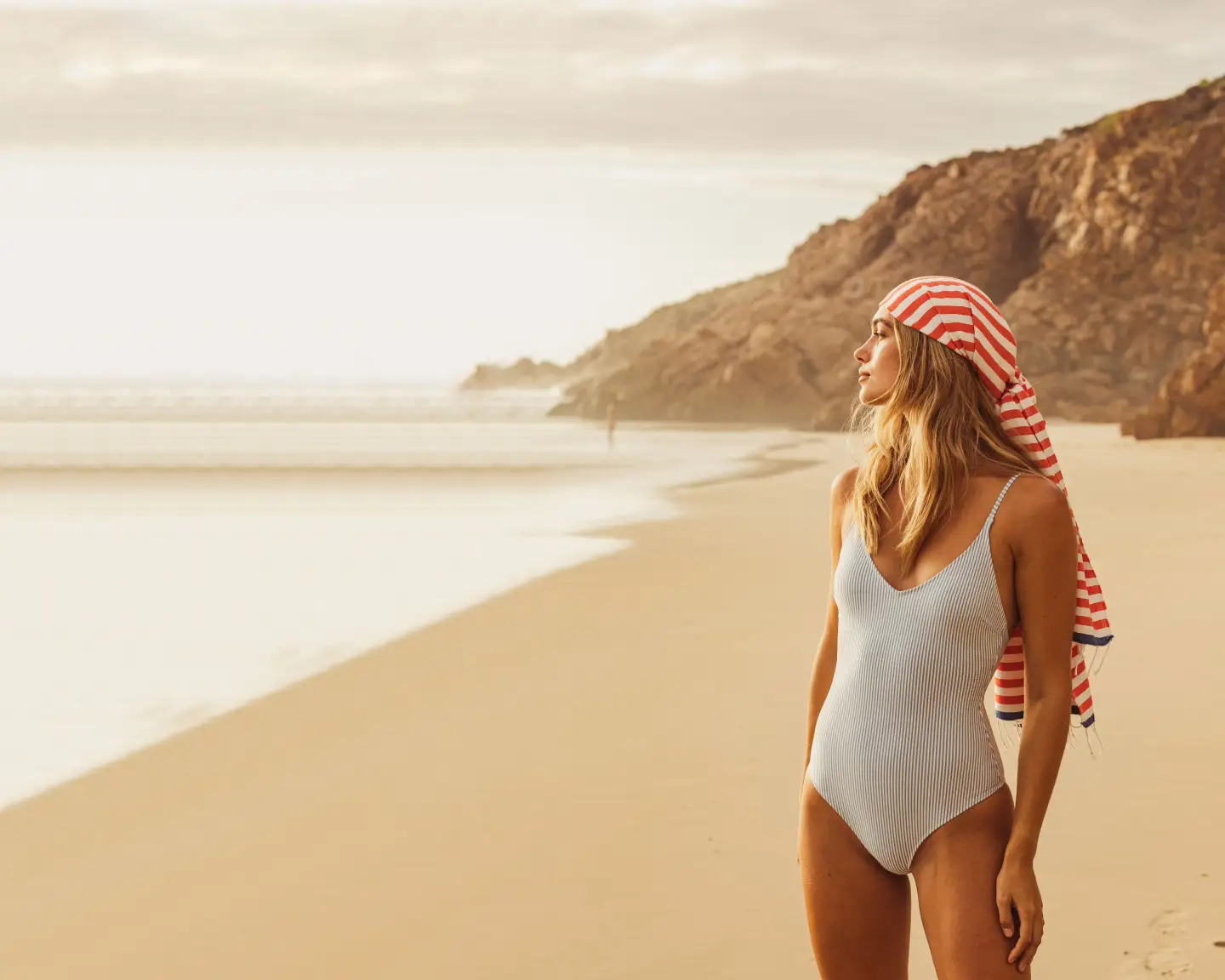 Mujer con bañador azul claro y sombrero de rayas mirando pensativamente al mar, de pie en la playa.