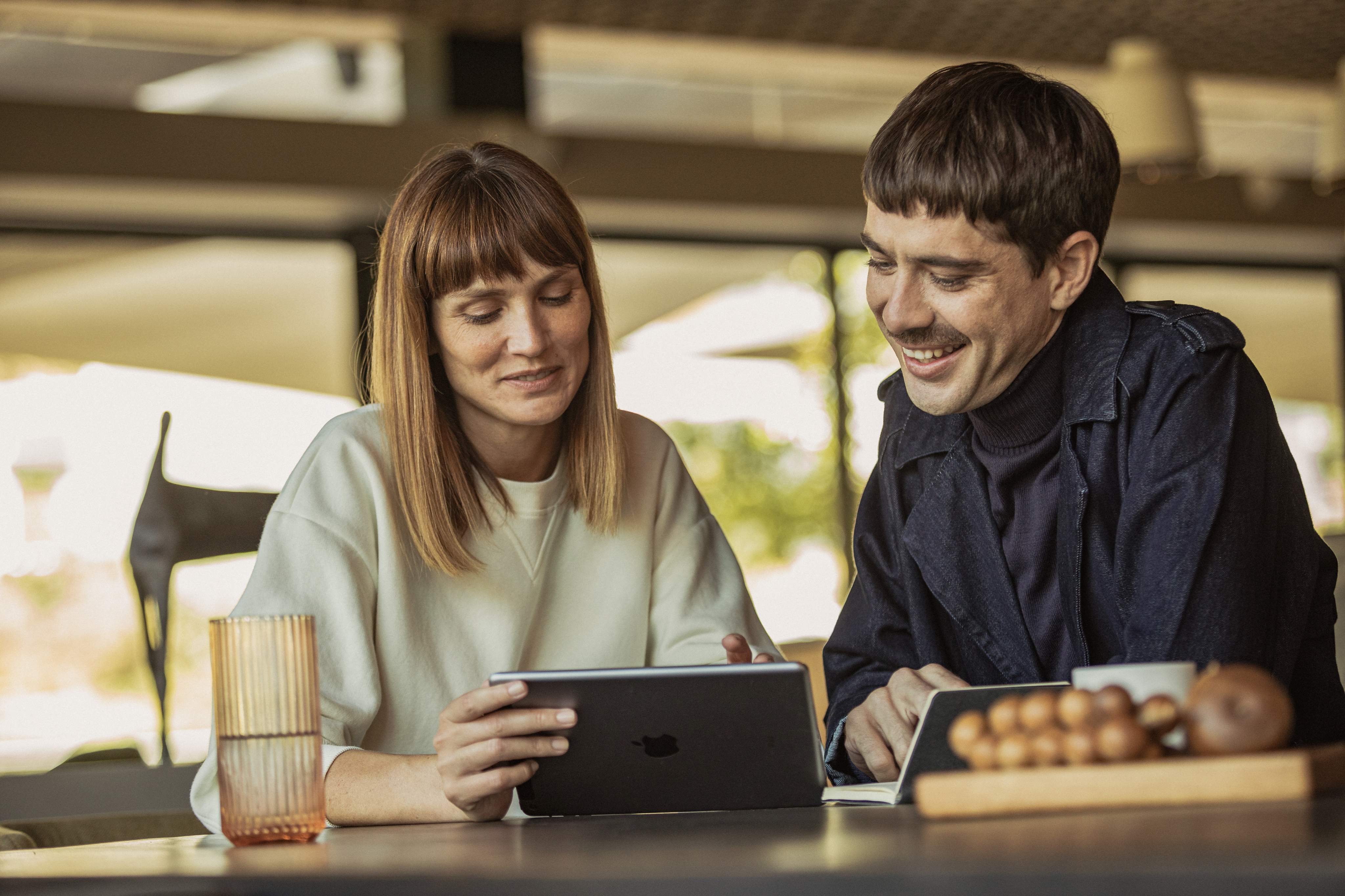 Egy fiatal férfi és egy fiatal nő ül egy asztalnál, és a tabletjüket nézik.