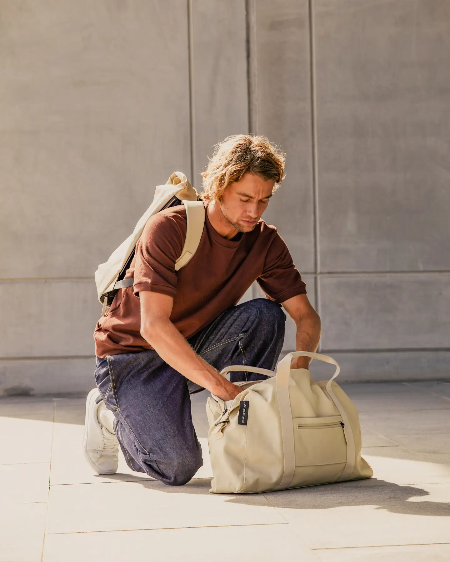 Un hombre con una camiseta marrón y unos vaqueros azules está arrodillado en el suelo y está buscando en una bolsa beige.