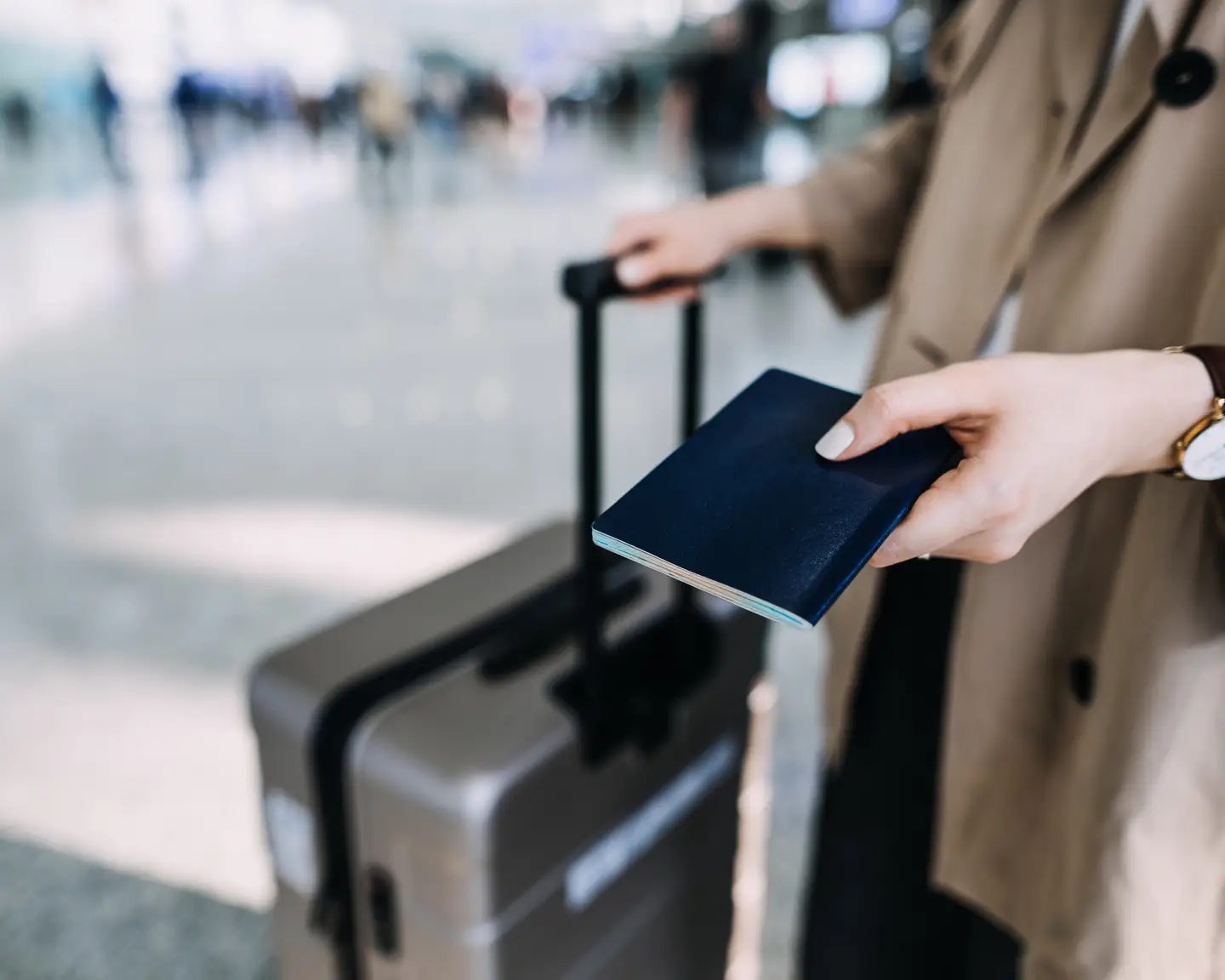 Primer plano de un pasaporte azul sostenido por un viajero con una maleta con ruedas en el aeropuerto.