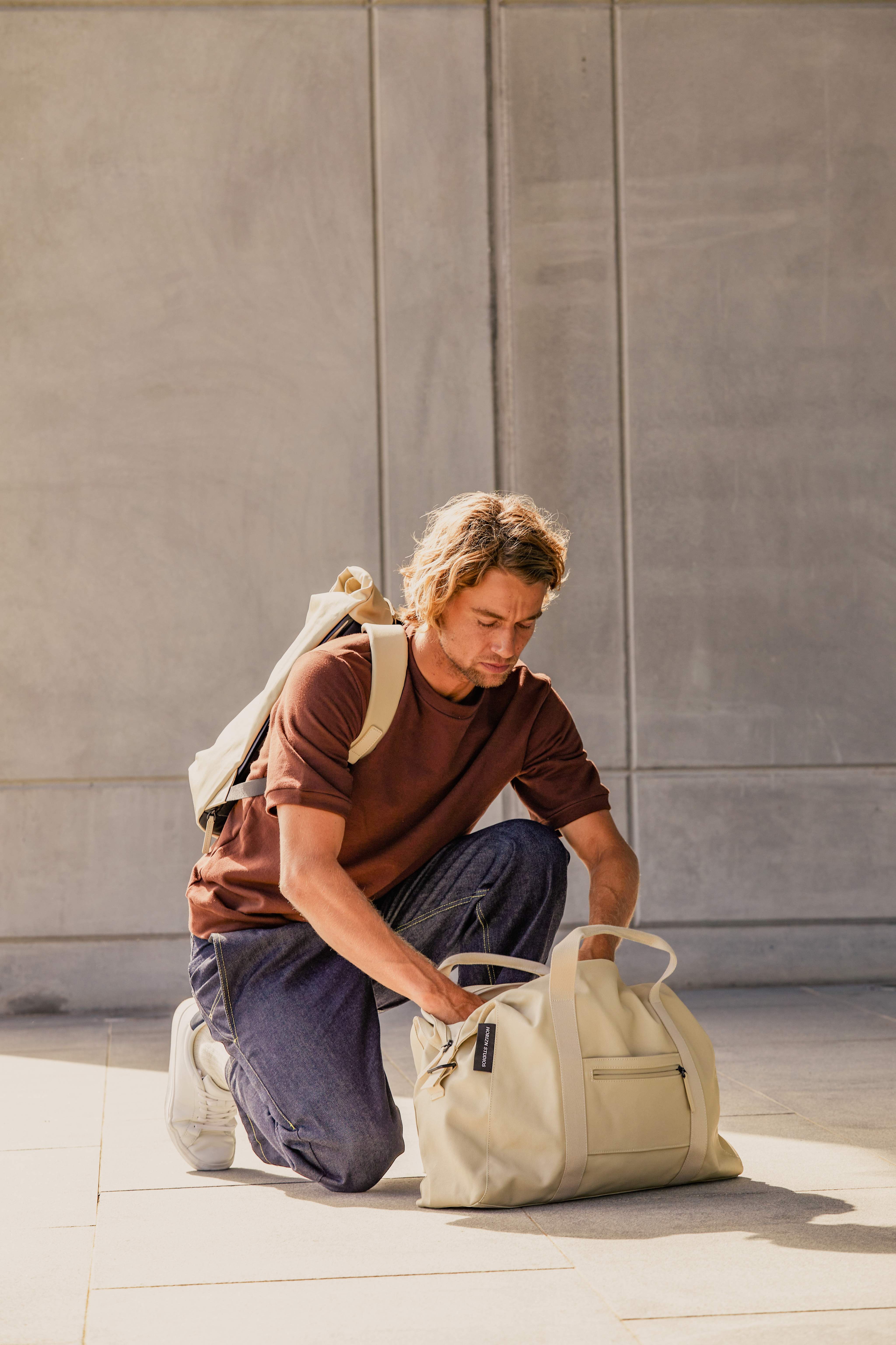 Un homme vêtu d'un t-shirt marron et d'un jean bleu est agenouillé sur le sol et fouille dans un sac beige.