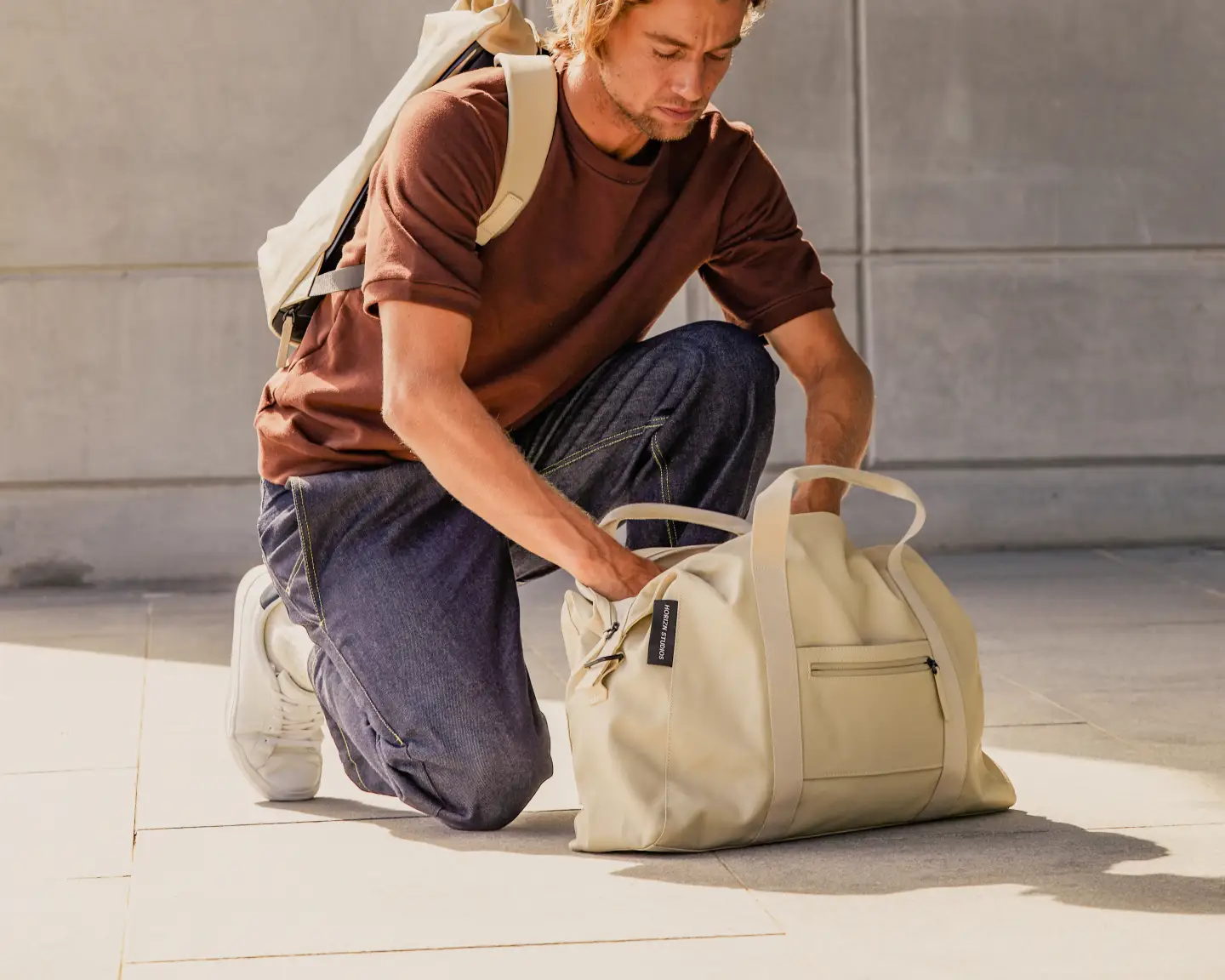 Un homme vêtu d'un t-shirt marron et d'un jean bleu est agenouillé sur le sol et fouille dans un sac beige.