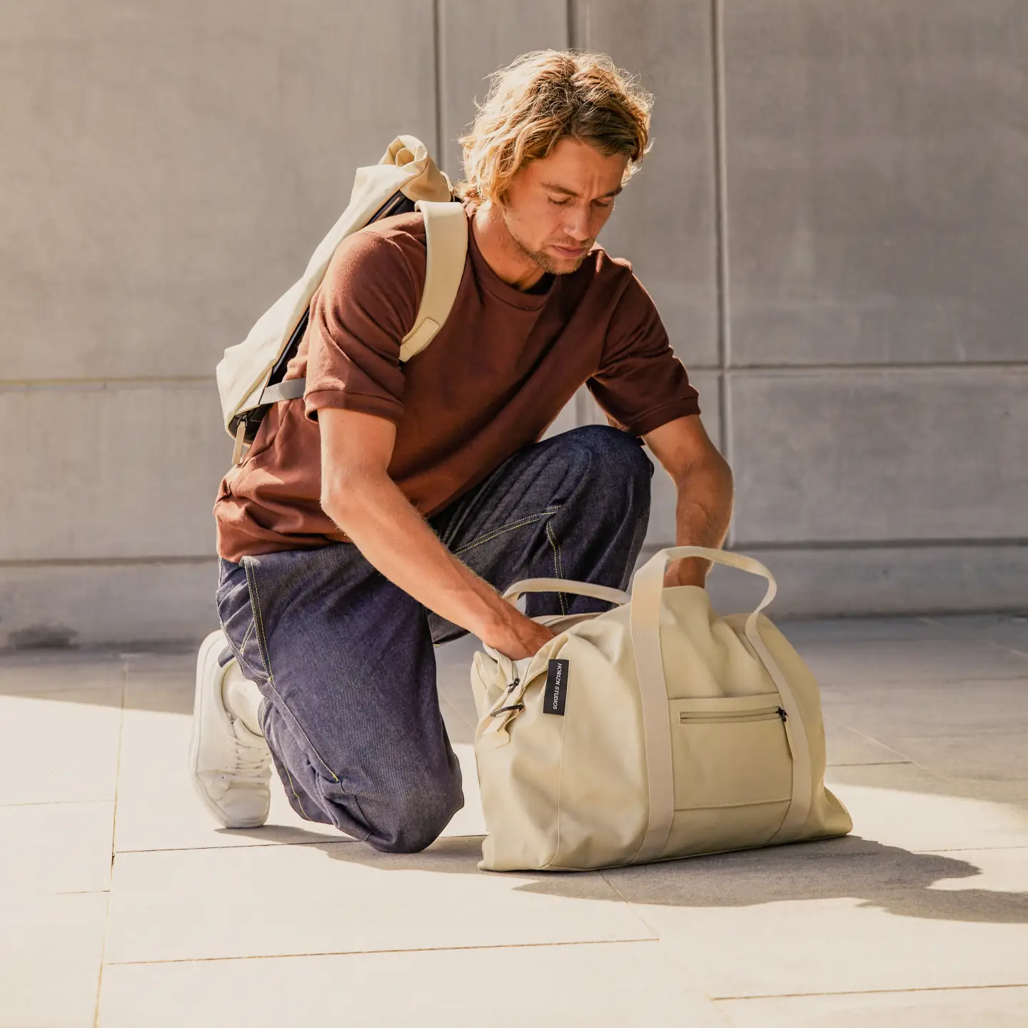 Un homme vêtu d'un t-shirt marron et d'un jean bleu est agenouillé sur le sol et fouille dans un sac beige.