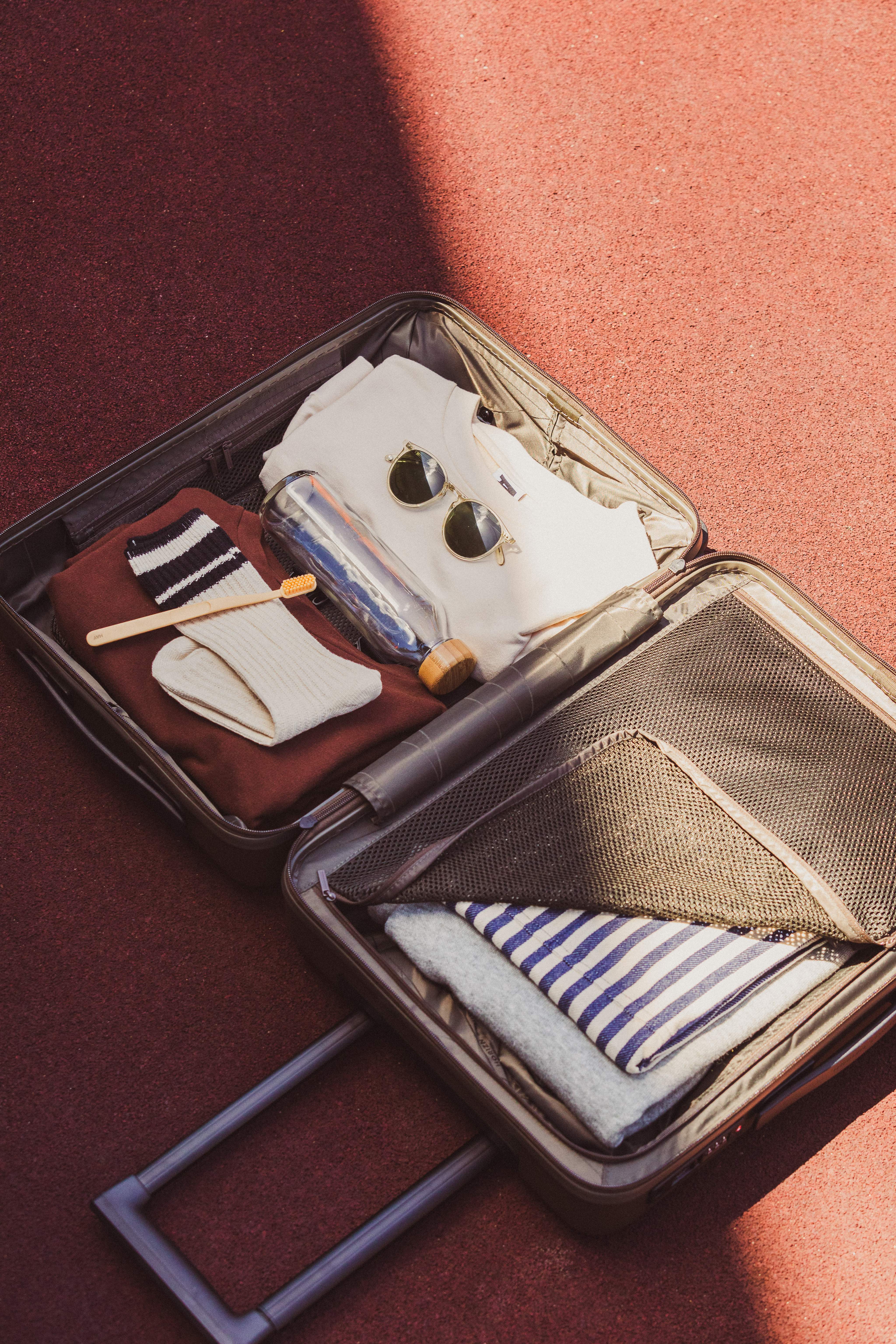 Une valise ouverte est posée sur le sol, contenant une brosse à dents, des chaussettes, des lunettes de soleil, une bouteille d’eau vide et quelques vêtements.