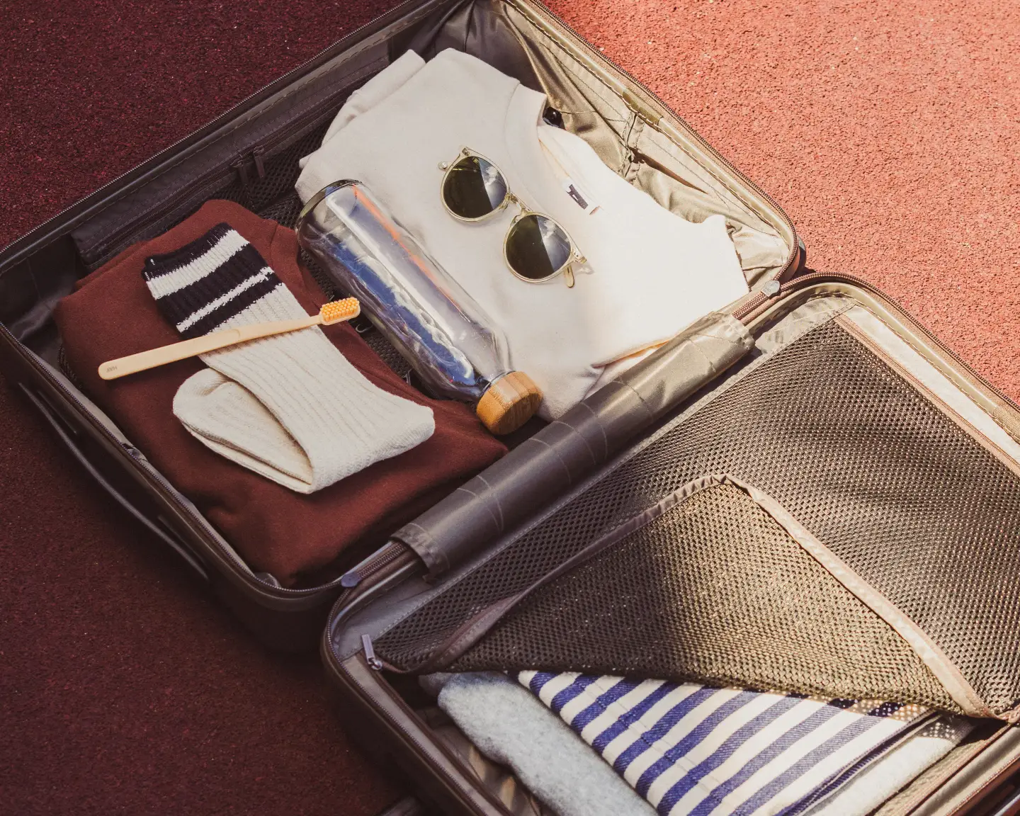 Une valise ouverte est posée sur le sol, contenant une brosse à dents, des chaussettes, des lunettes de soleil, une bouteille d’eau vide et quelques vêtements.