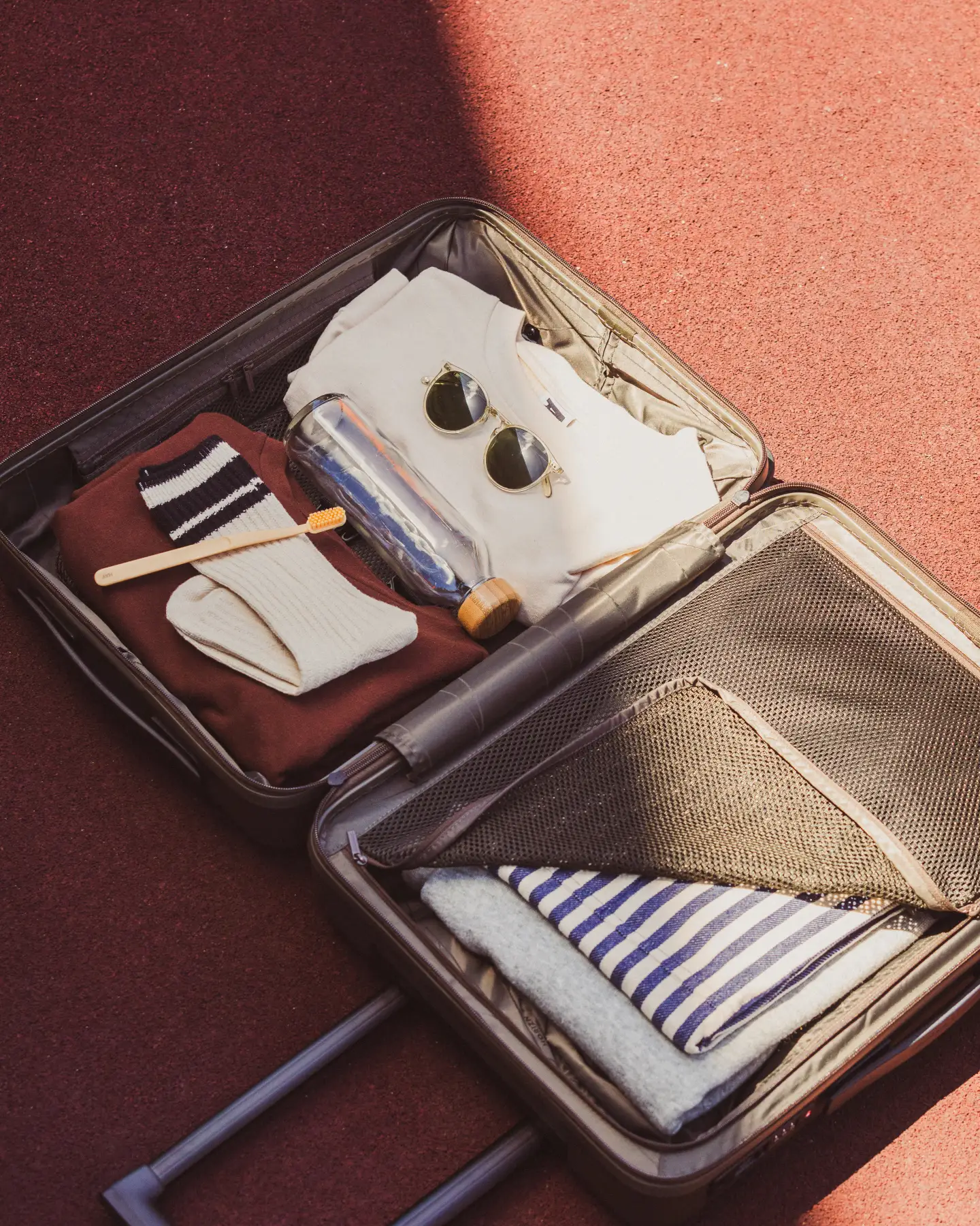 Une valise ouverte est posée sur le sol, contenant une brosse à dents, des chaussettes, des lunettes de soleil, une bouteille d’eau vide et quelques vêtements.