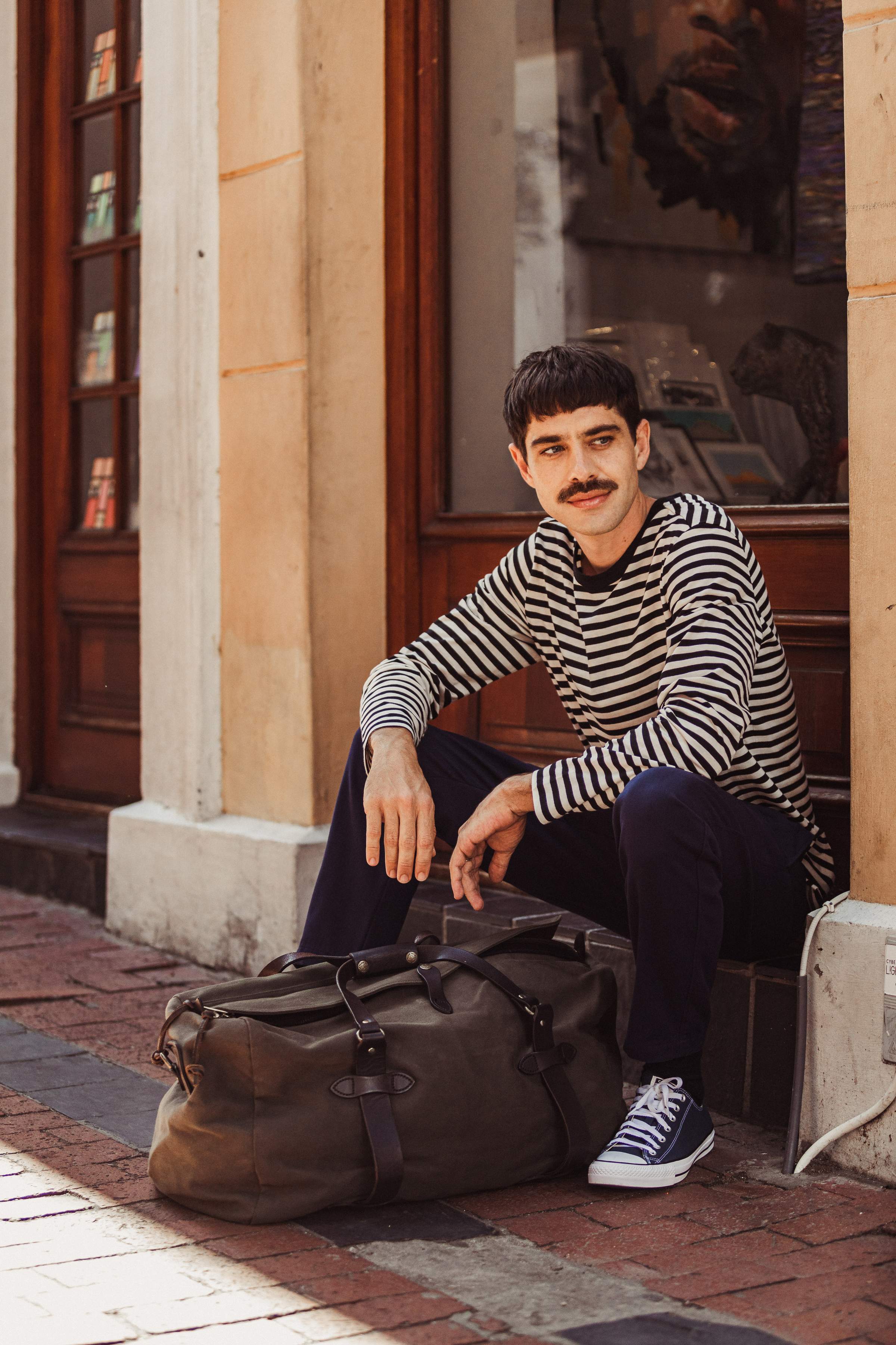 Hombre con camisa de rayas esperando con su equipaje frente a un edificio de ladrillo.
