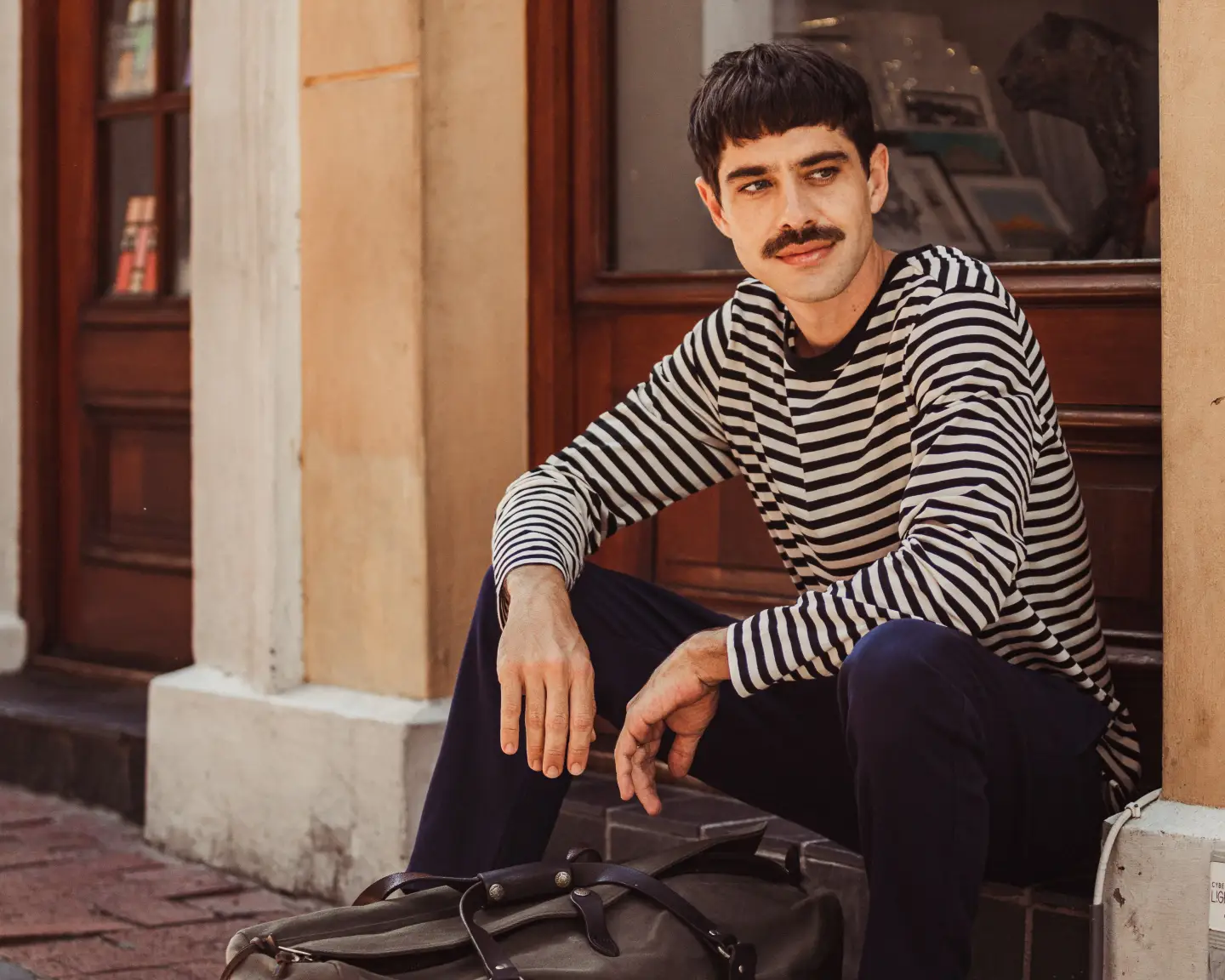 A man in a striped shirt waits with his baggage in front of a brick building.