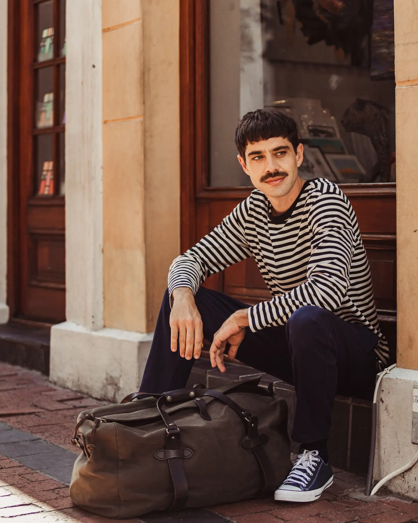 Hombre con camisa de rayas esperando con su equipaje frente a un edificio de ladrillo.