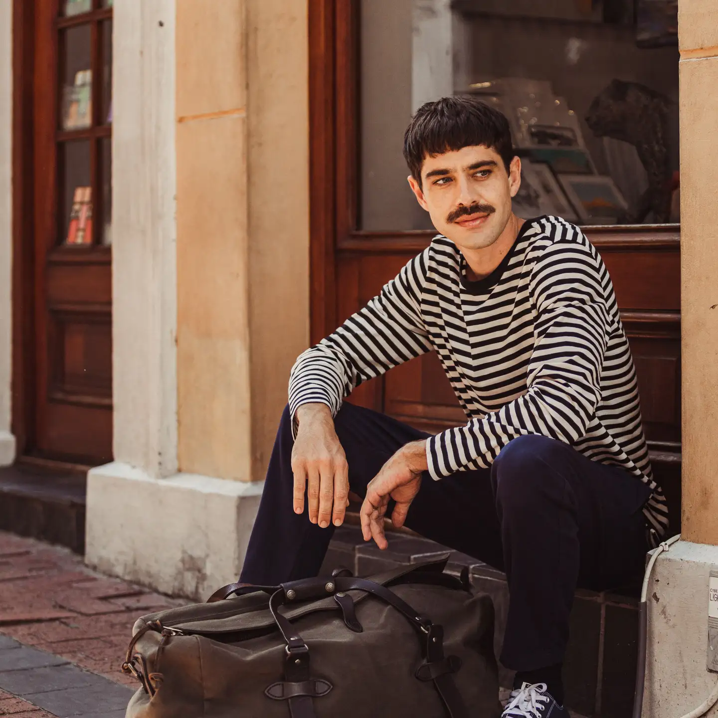 Hombre con camisa de rayas esperando con su equipaje frente a un edificio de ladrillo.