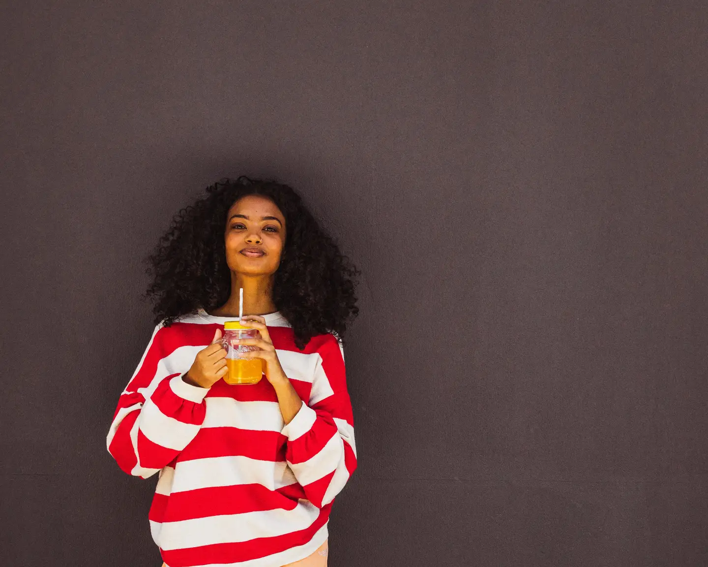Una mujer está apoyada contra una pared, lleva un suéter a rayas y sostiene un frasco de zumo.