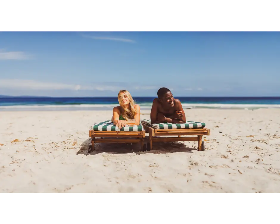 Zwei Reisende entspannen auf Sonnenliegen an einem weißen Sandstrand mit Blick auf das Meer.