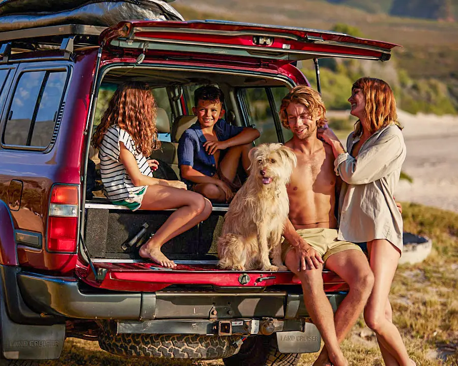 Una familia en la playa con un coche rojo grande. Dos niños y un hombre están sentados juntos con un perro en el maletero del coche, la mujer está de pie junto a ellos.